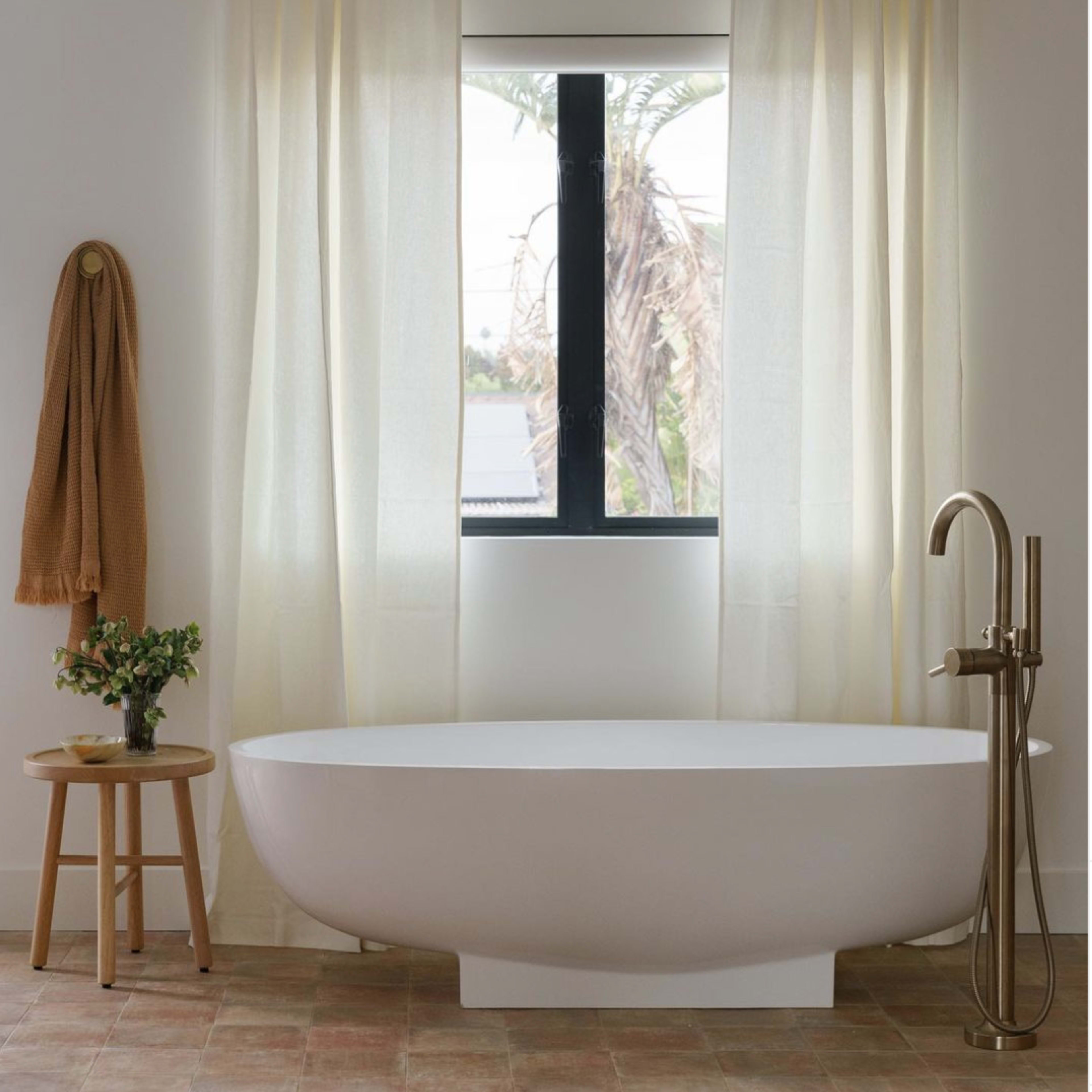 A modern bathroom features a freestanding white bathtub beside a small wooden stool, with sheer curtains framing a window overlooking palm trees.
