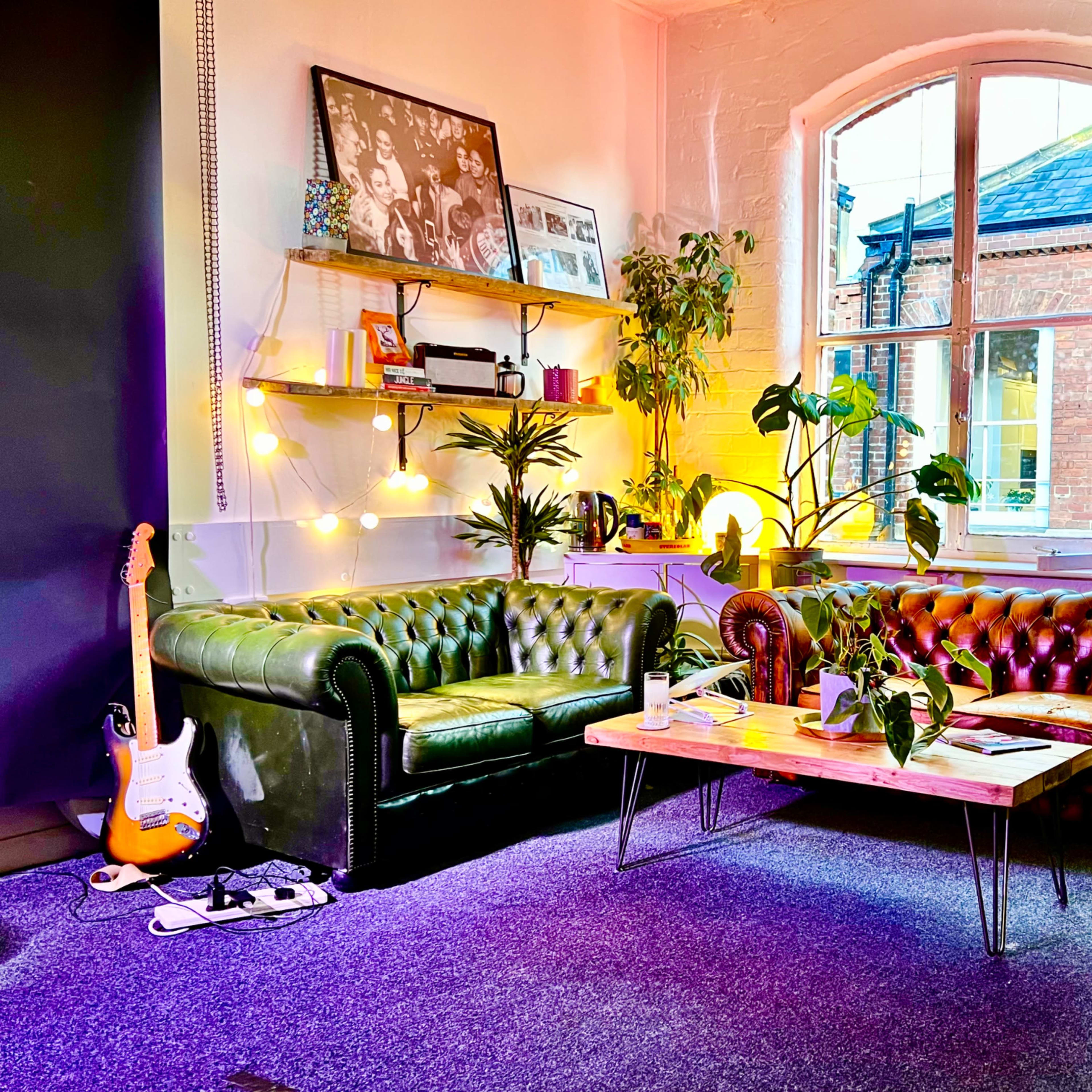 The scene features a cozy seating area with two leather sofas, a wooden coffee table surrounded by plants, and a large window providing natural light.