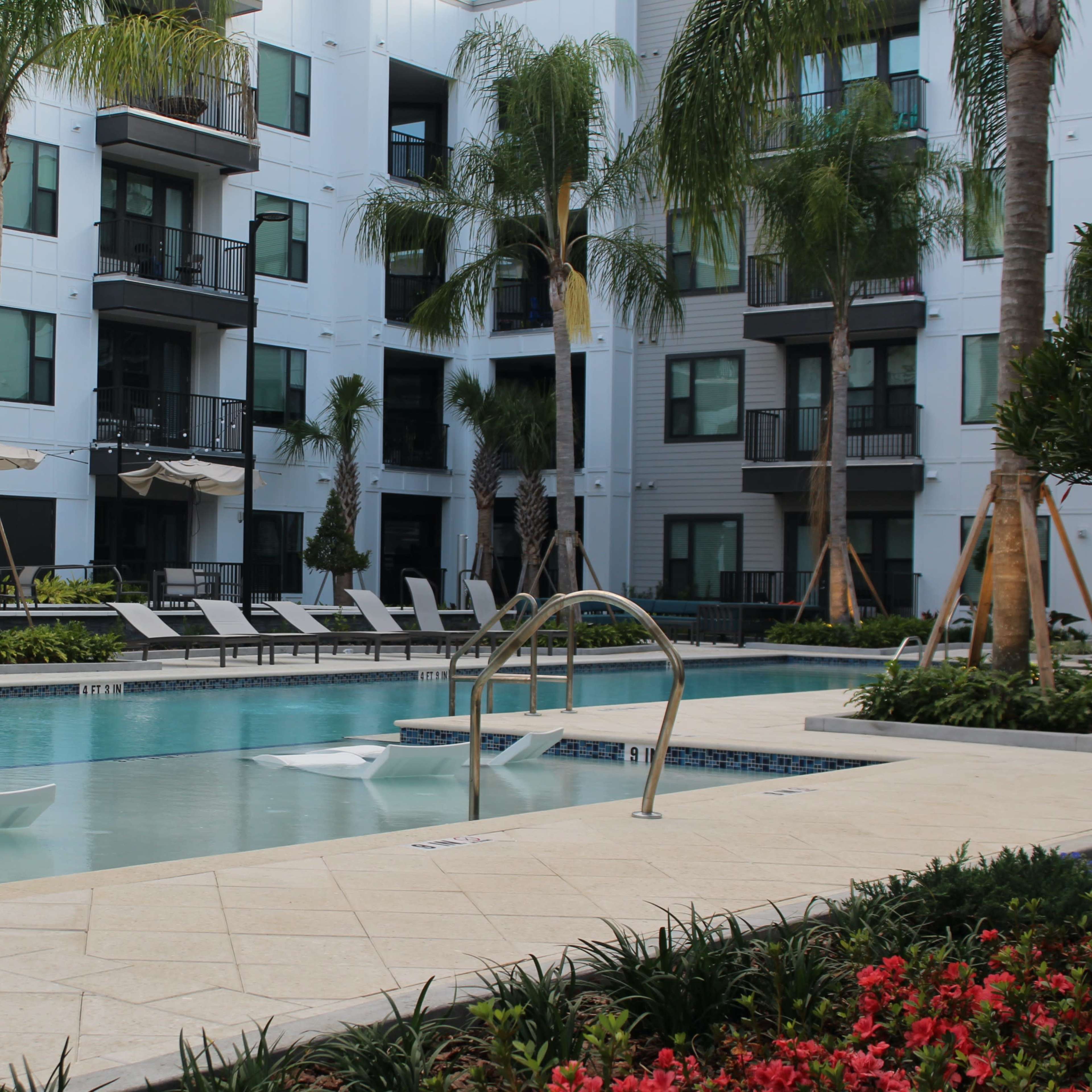 A swimming pool surrounded by palm trees and lounge chairs is situated in a courtyard of a modern apartment complex.