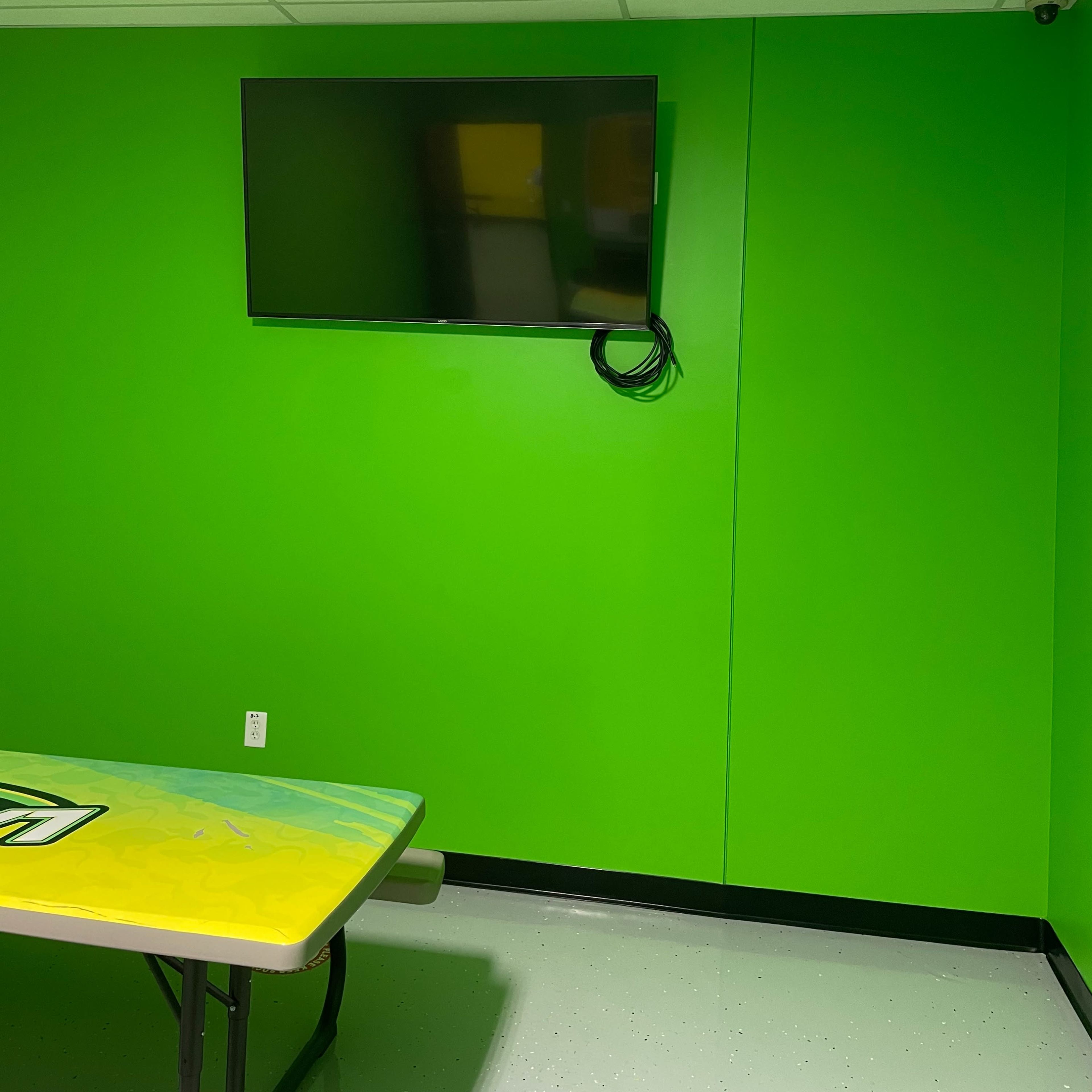 A green-walled room with a mounted flat-screen TV and a table covered with a colorful cloth.