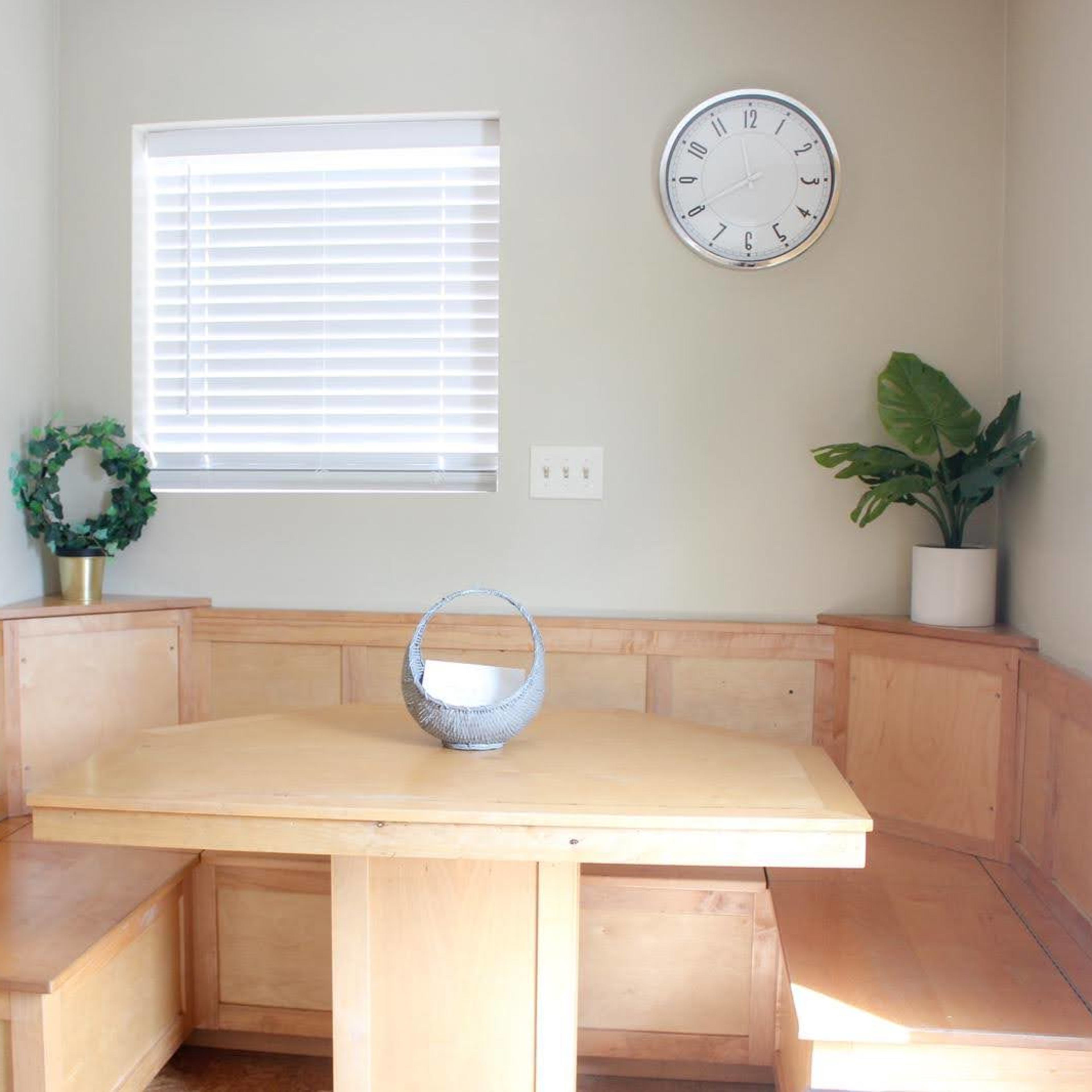 A corner dining nook with wooden seating, a round table, a decorative basket, and two plants next to a window.