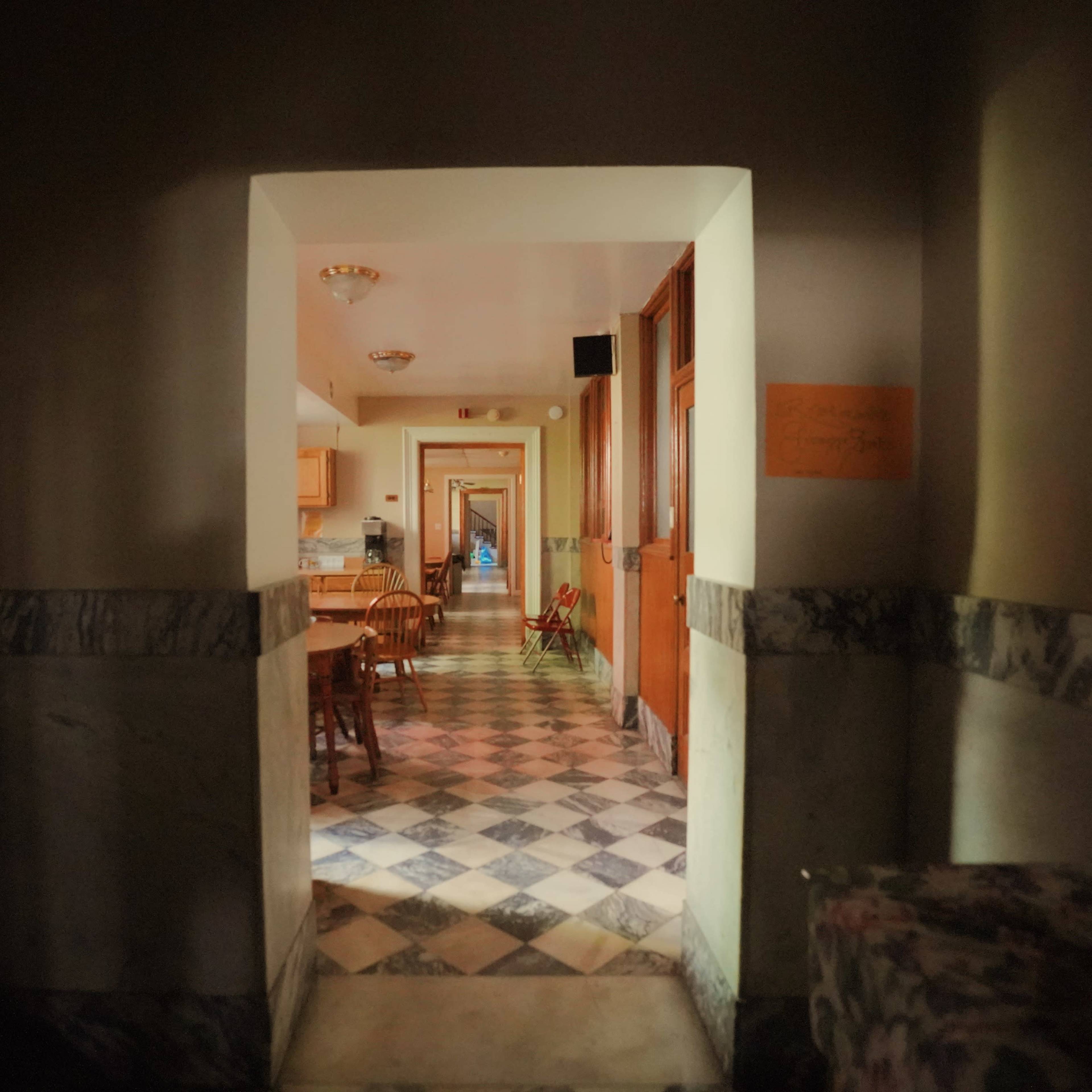 A hallway with a checkered floor leads to a series of doorways and a kitchen area at the far end.