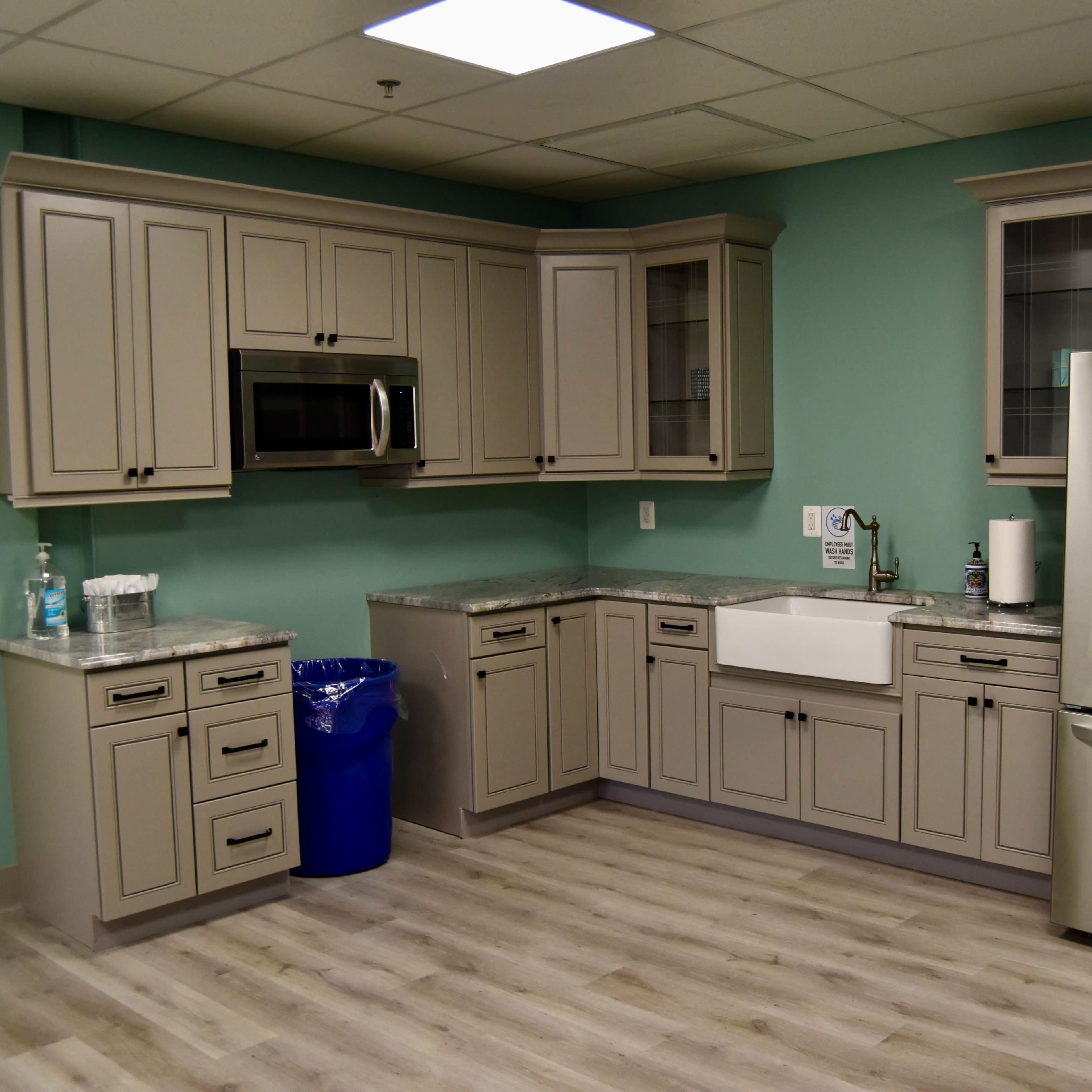 A kitchen featuring gray cabinetry, a stainless steel refrigerator, a large white sink, and light wood flooring against a teal wall.