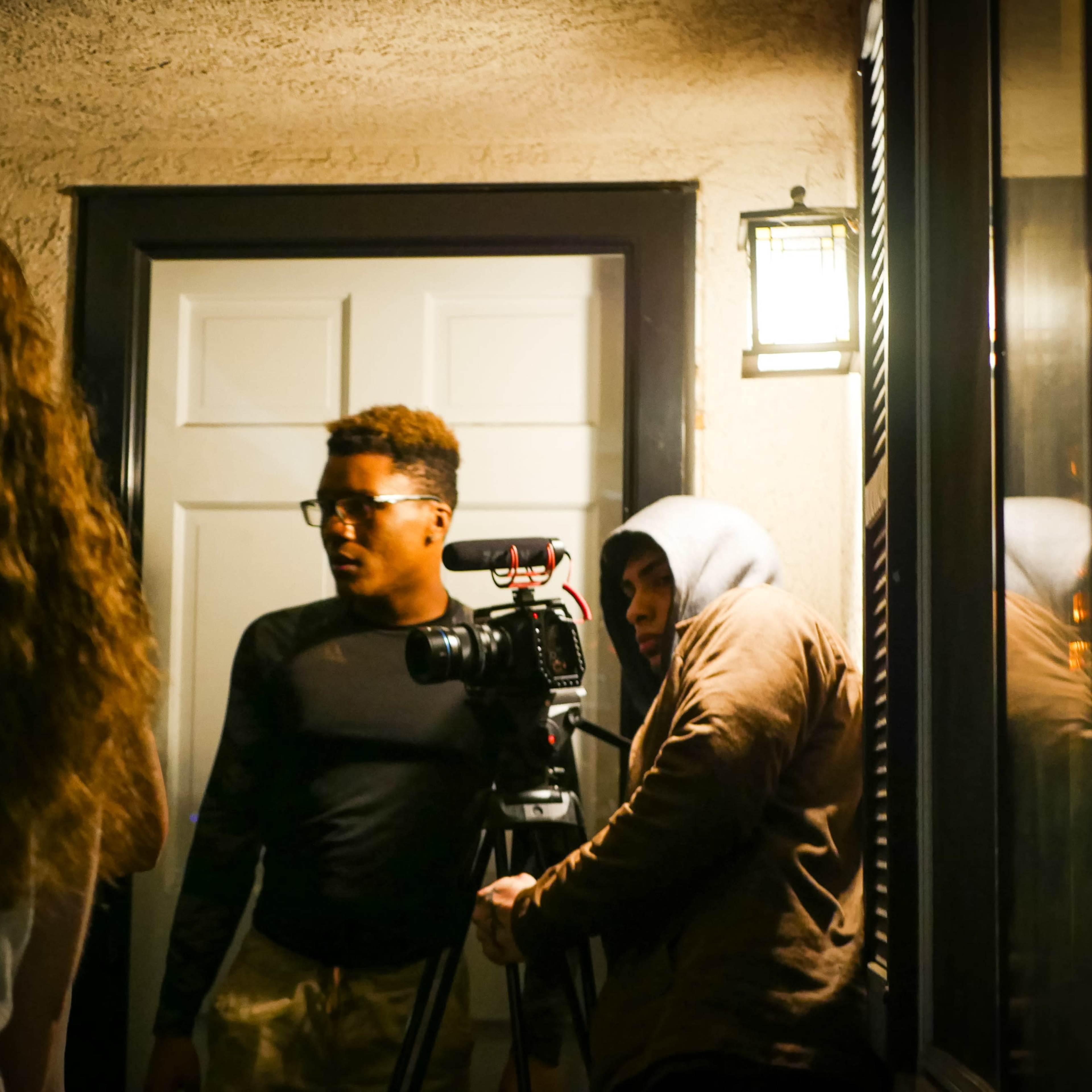 A group of three people is gathered near a doorway, with one person filming using a camera on a tripod while another wears a hoodie.