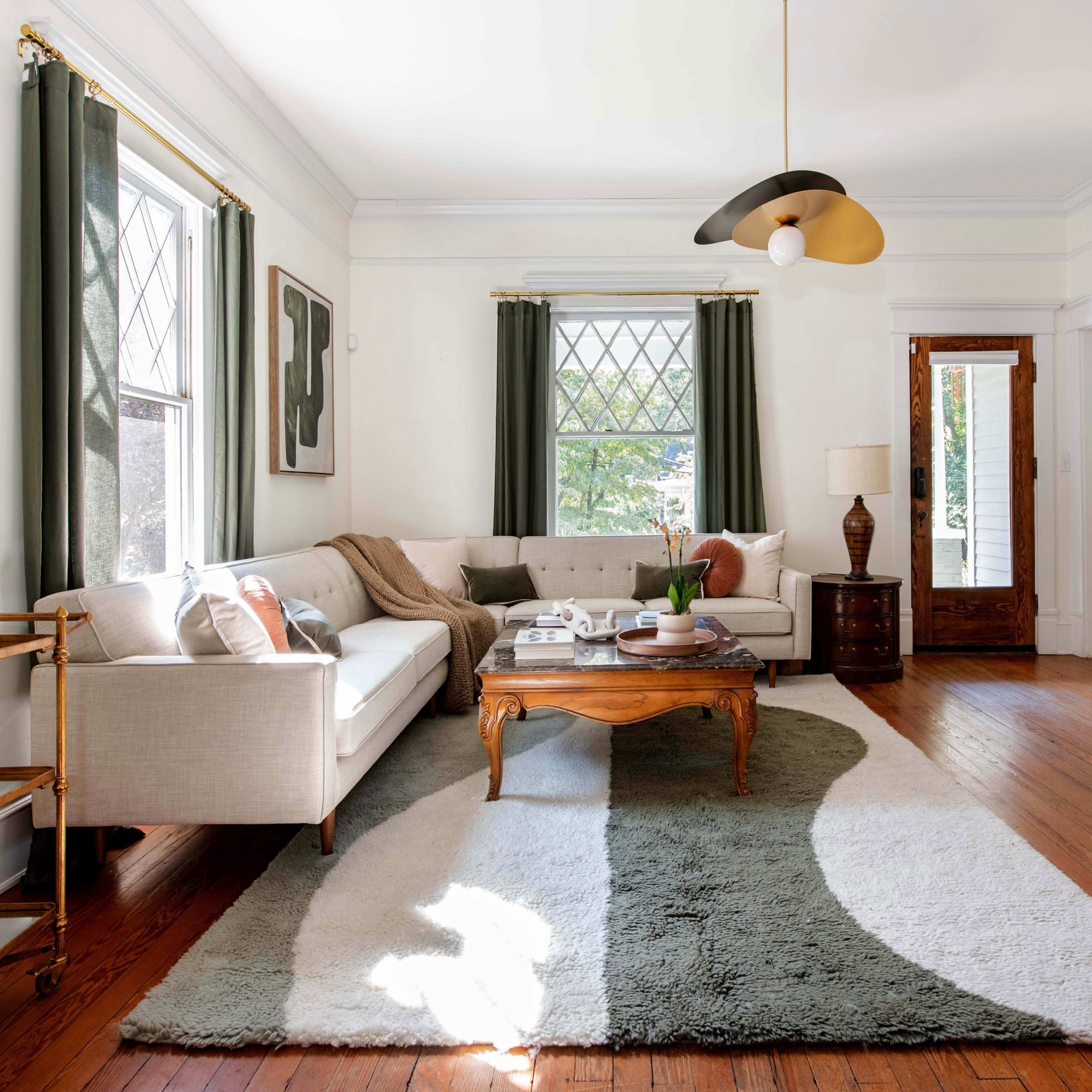 A bright living room features a beige sofa, a round coffee table, and a large area rug, complemented by green curtains and hardwood floors.
