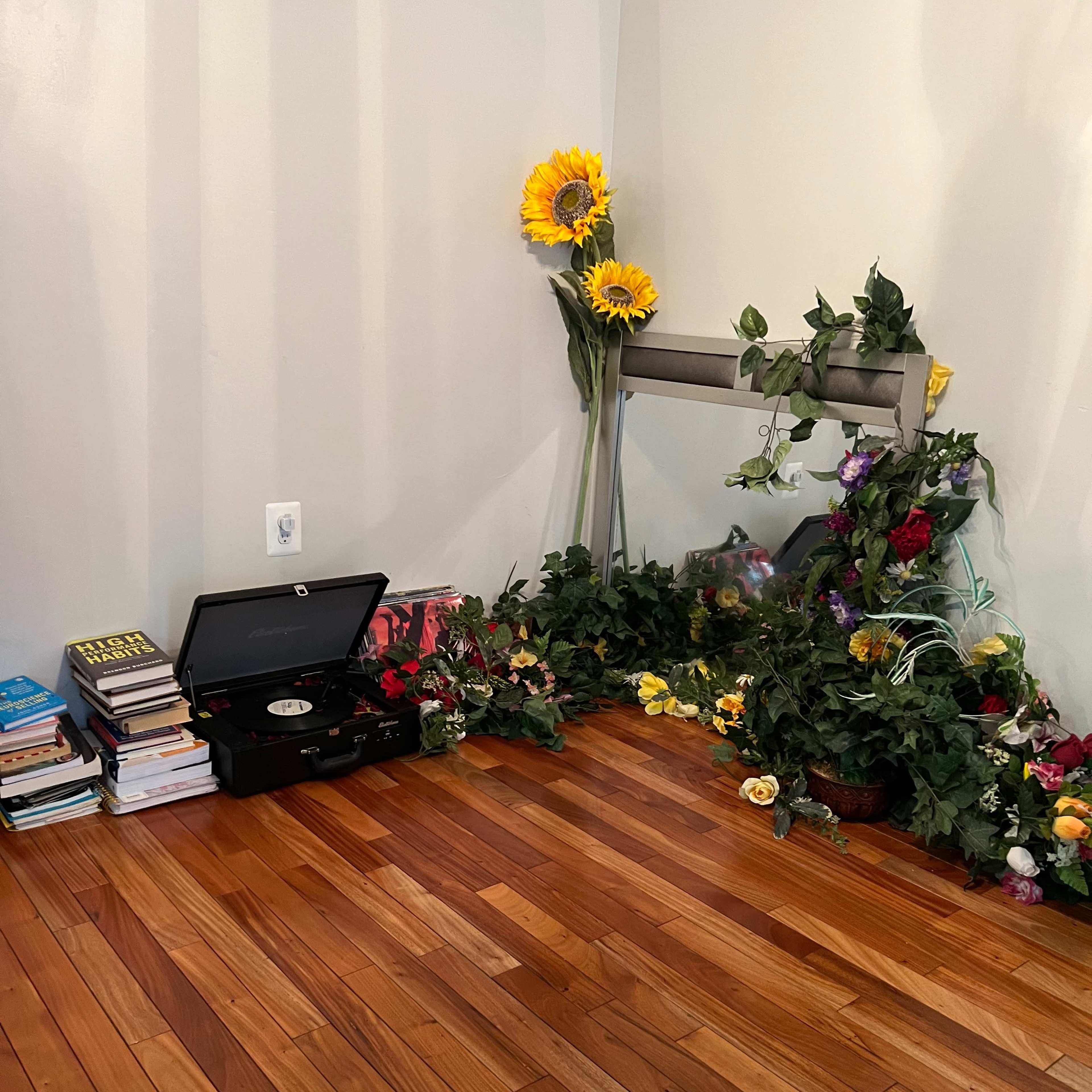 A corner of a room features a stack of books, a record player, and an arrangement of artificial flowers and greenery.