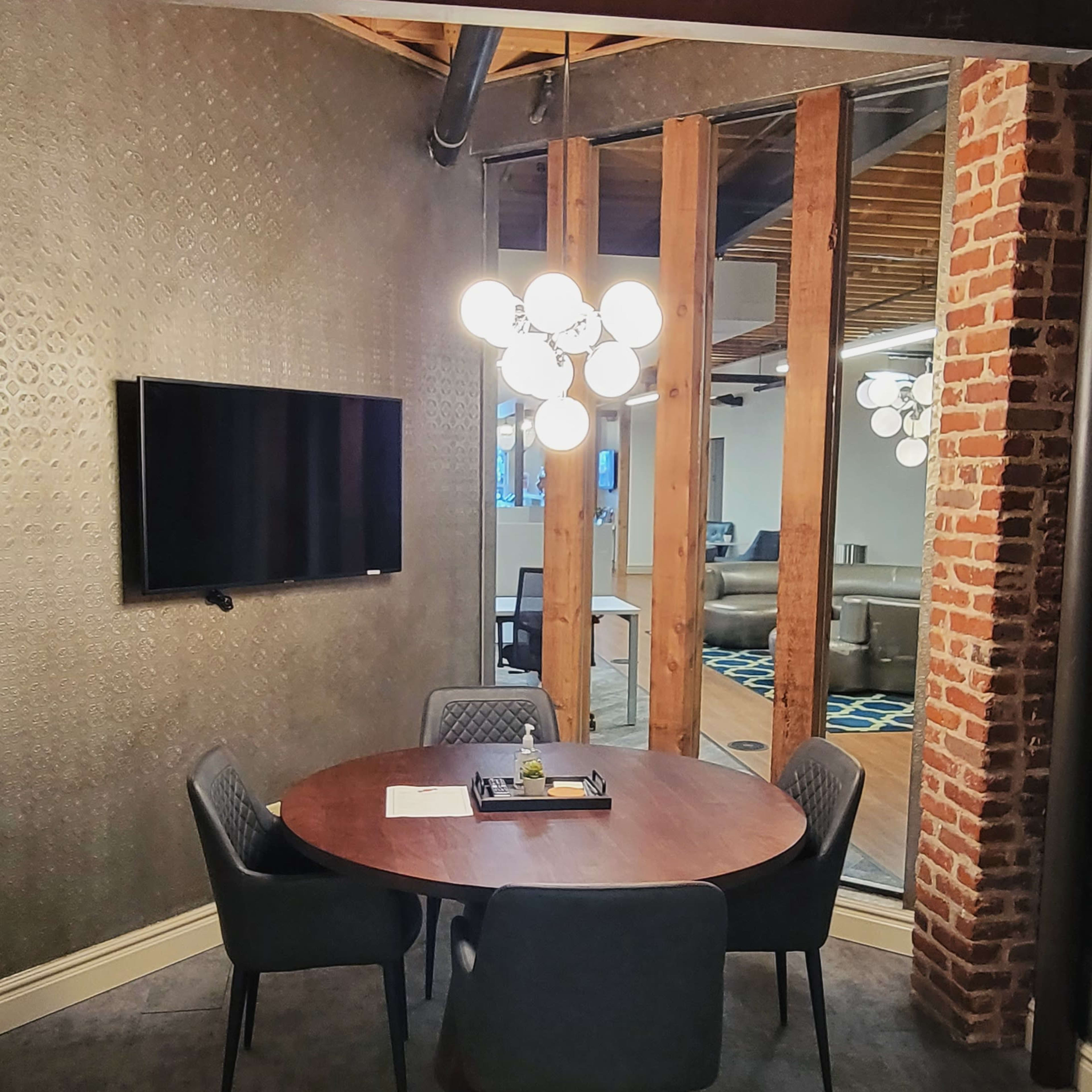 A small conference room features a round wooden table with four chairs, a television mounted on the wall, and a decorative light fixture.