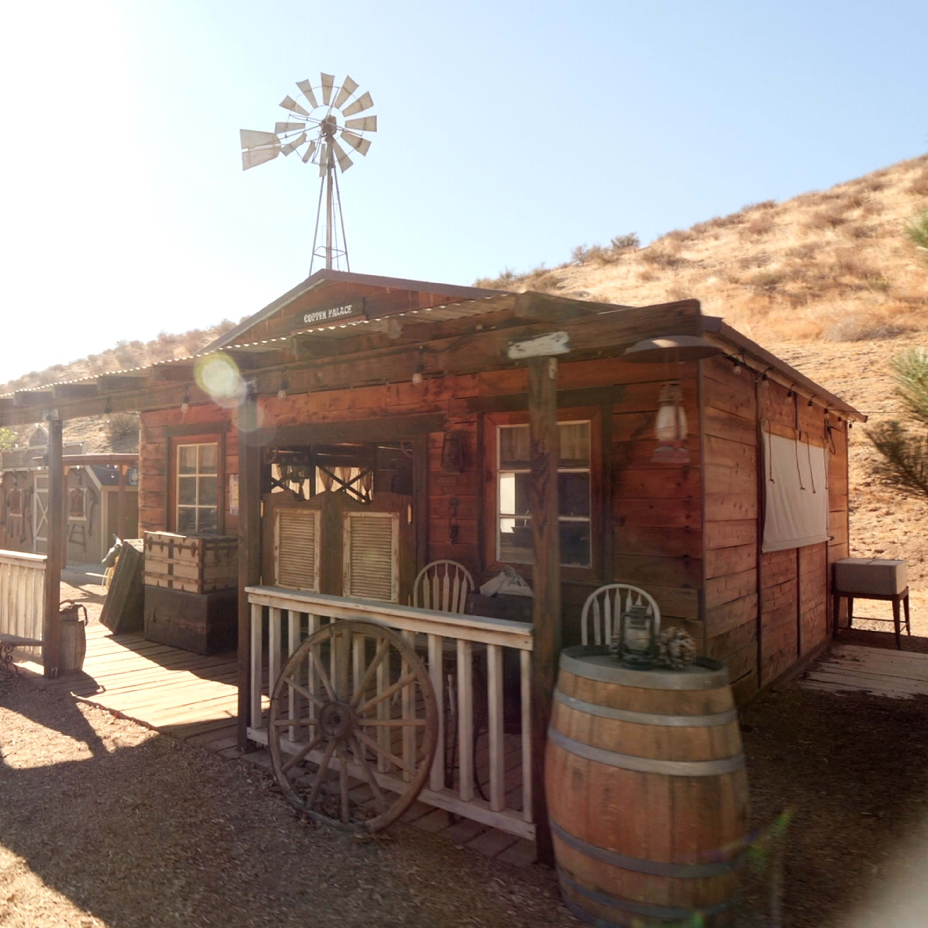 A rustic wooden building with a covered porch and a windmill stands in a dry landscape.