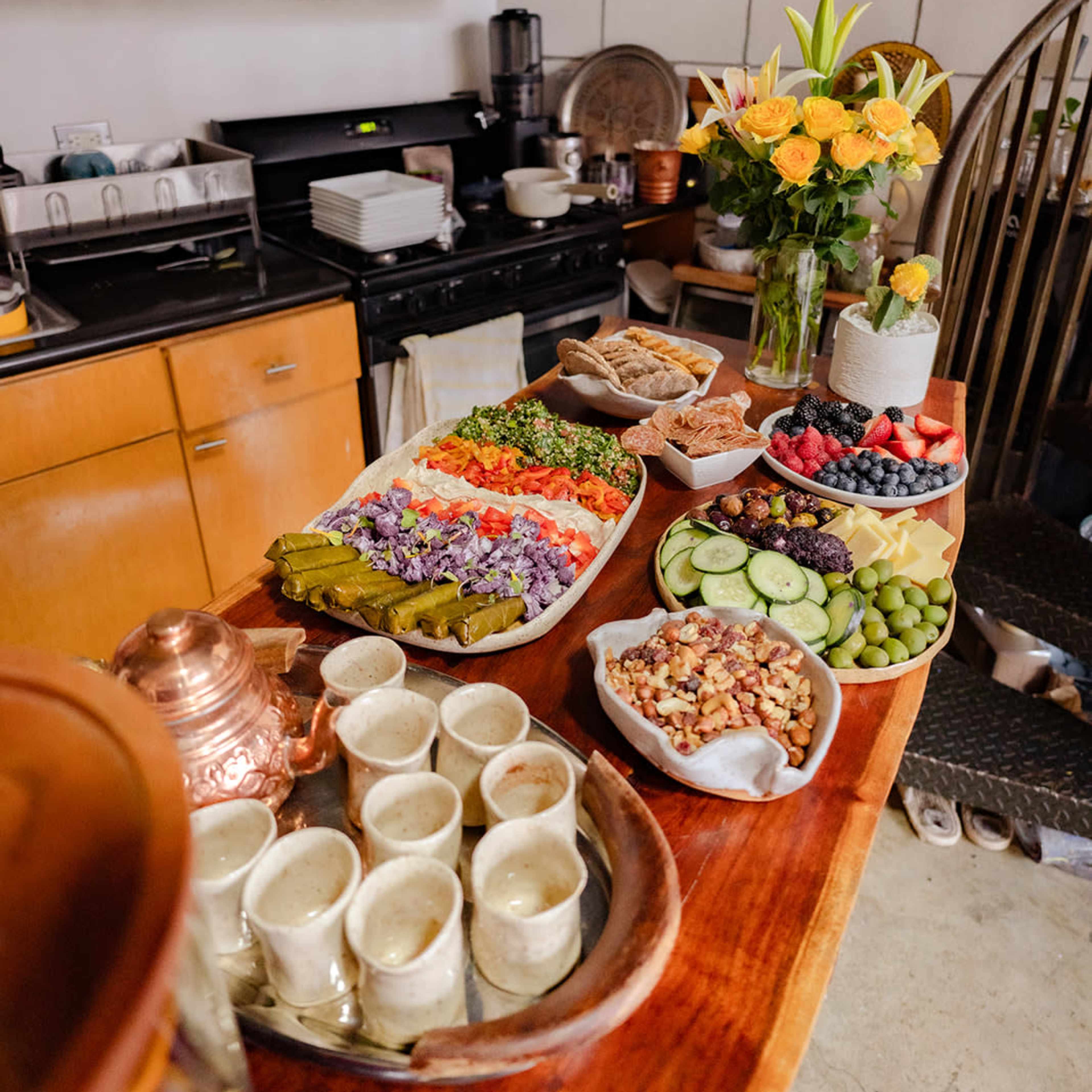 A dining table displays a variety of food items, including salads, fruits, and snacks, alongside a vase of flowers in a kitchen setting.