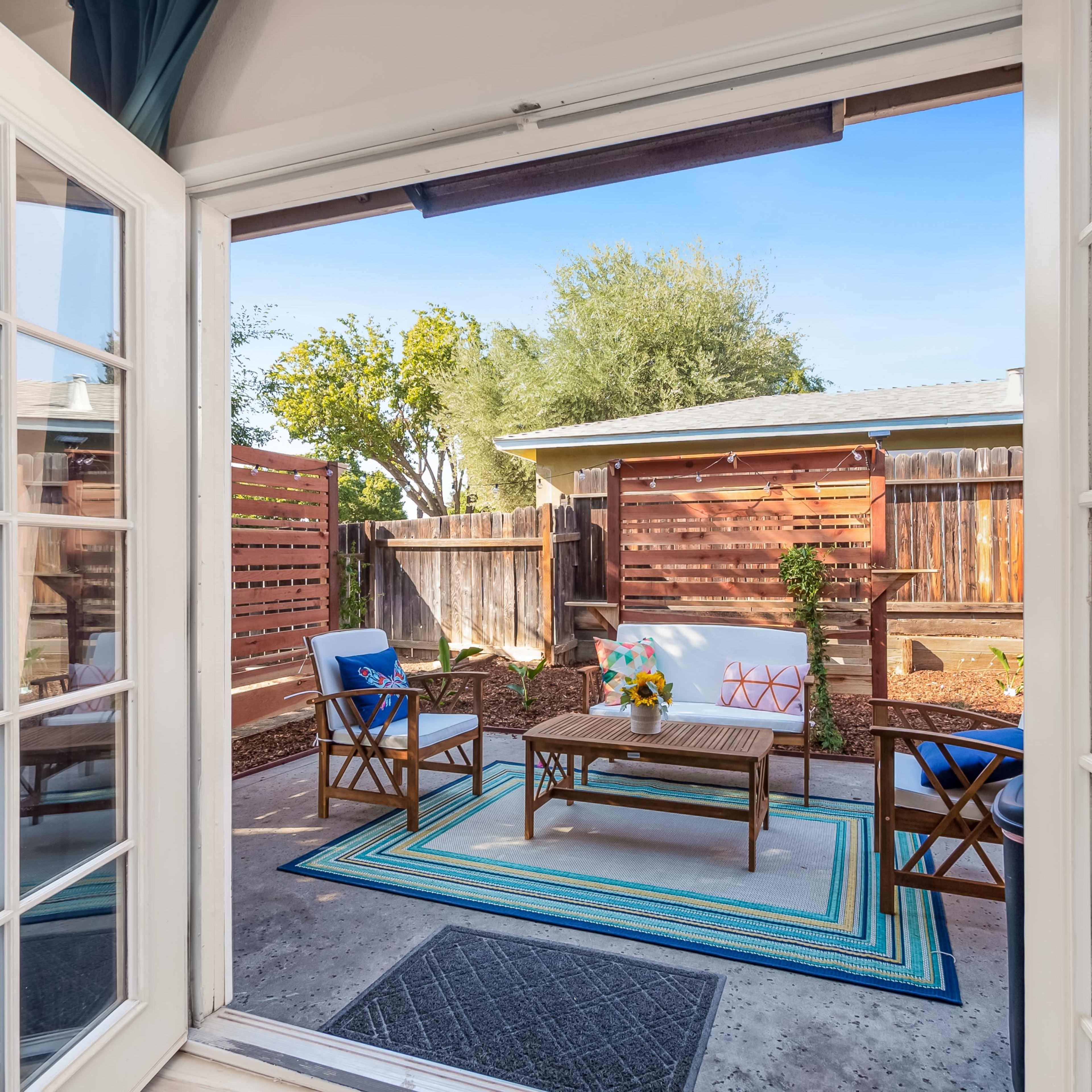 A view through double doors reveals a patio area with a seating arrangement, colorful rug, and landscaped garden.