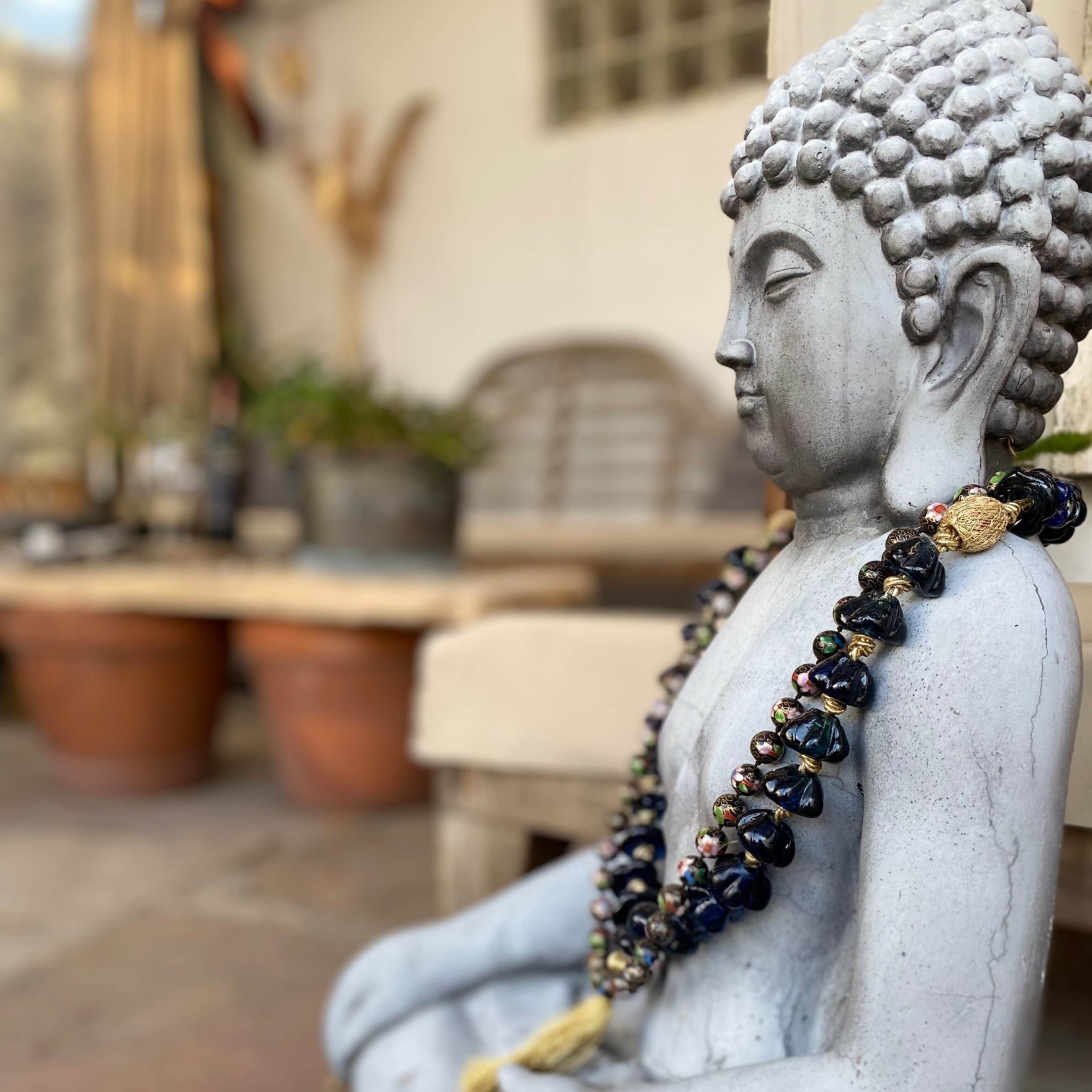 A gray stone Buddha statue sits with a beaded necklace draped around its neck in a garden setting with potted plants and wooden furniture in the background.