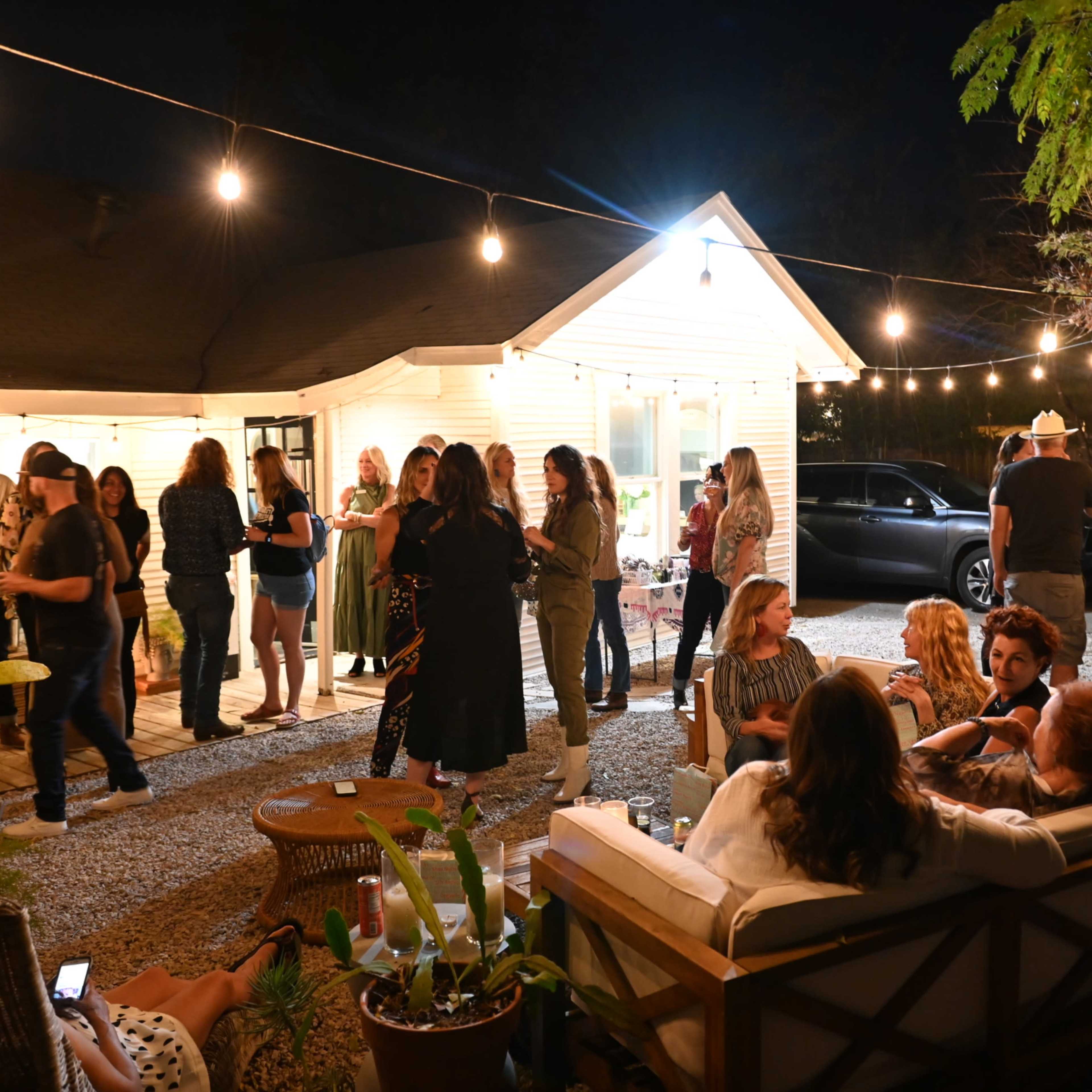 A group of people gathers at night in a backyard setting illuminated by string lights, with a house and a car visible in the background.