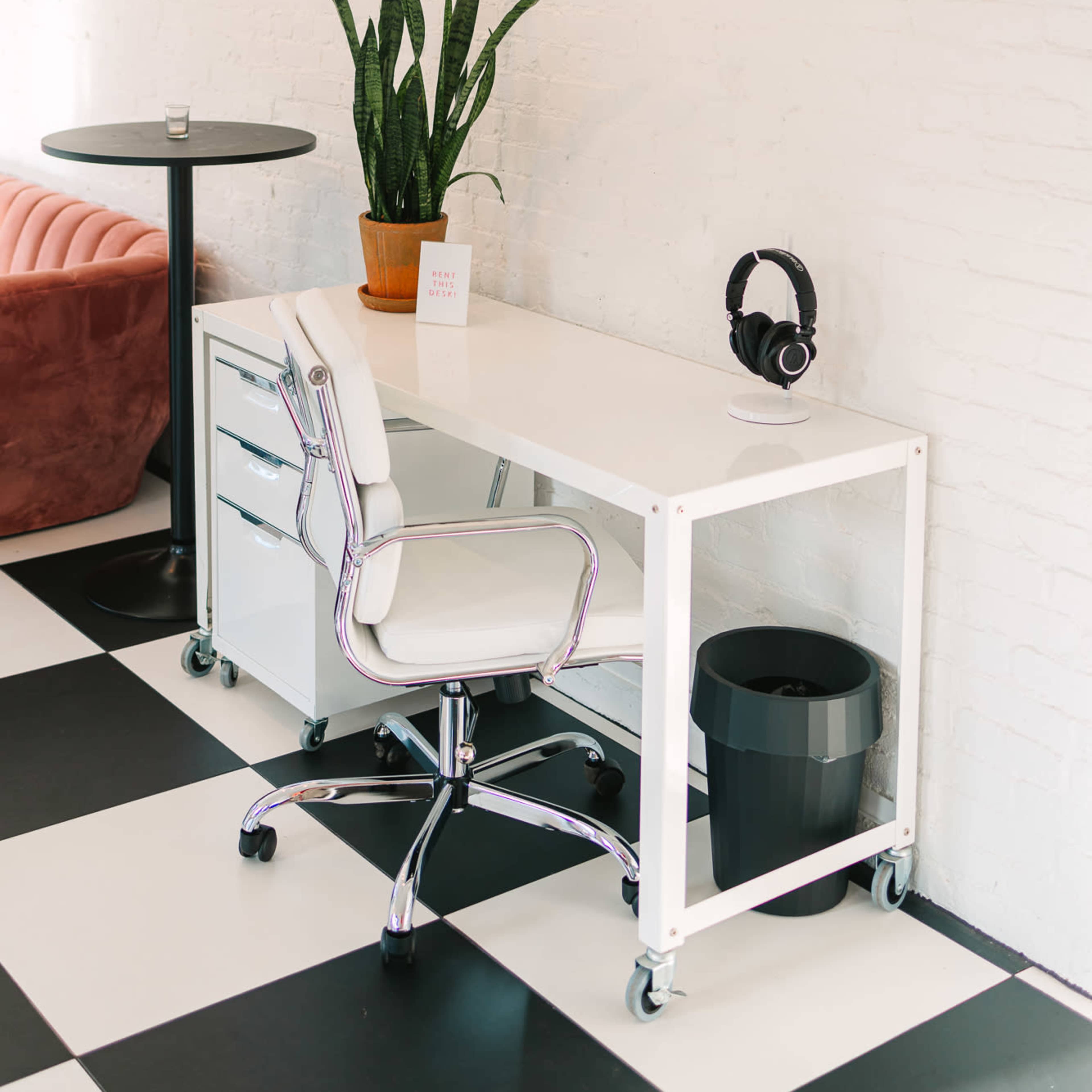A minimalist workspace features a white desk on wheels, a rolling chair, headphones, a potted plant, and a black trash can, all set against a checkered floor and a light-colored wall.