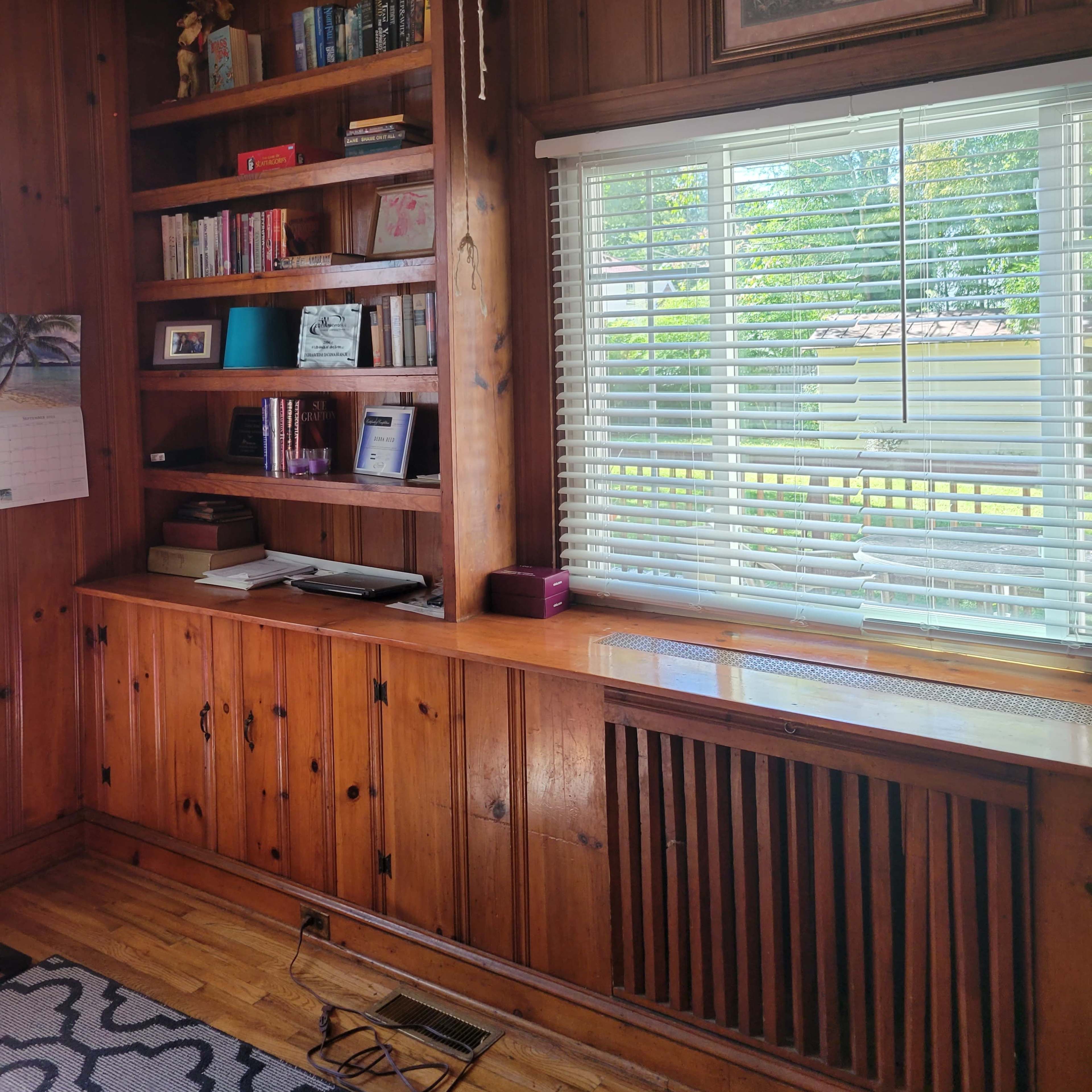 A wooden-paneled room featuring a bookshelf filled with books, a window with blinds, and a wall-mounted picture above a wooden shelf.
