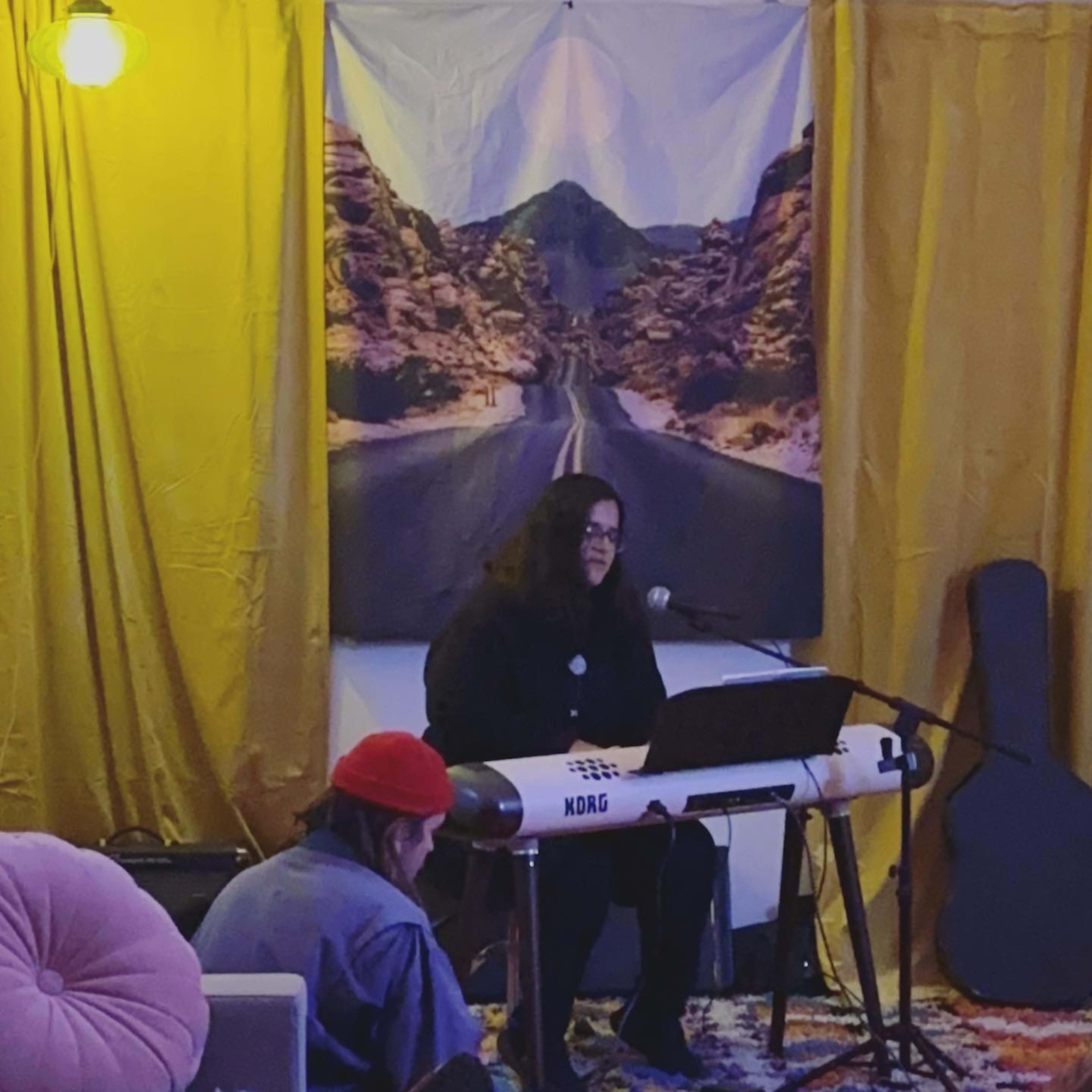 A musician sits at a keyboard on a stage in a cozy indoor setting, with a backdrop depicting a desert road and audience members seated in front.