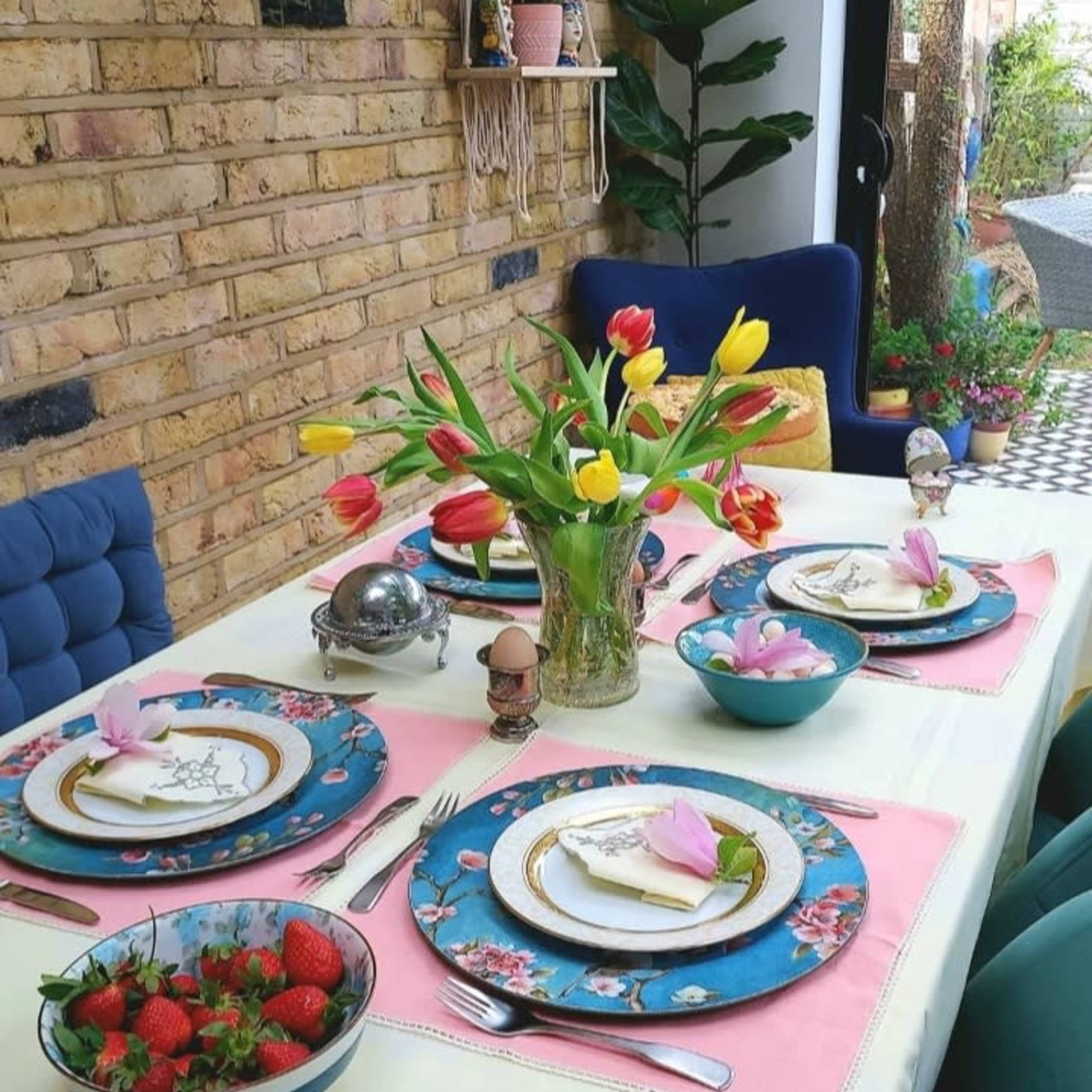 A dining table is elegantly set with colorful plates, fresh flowers in a vase, and bowls of strawberries.