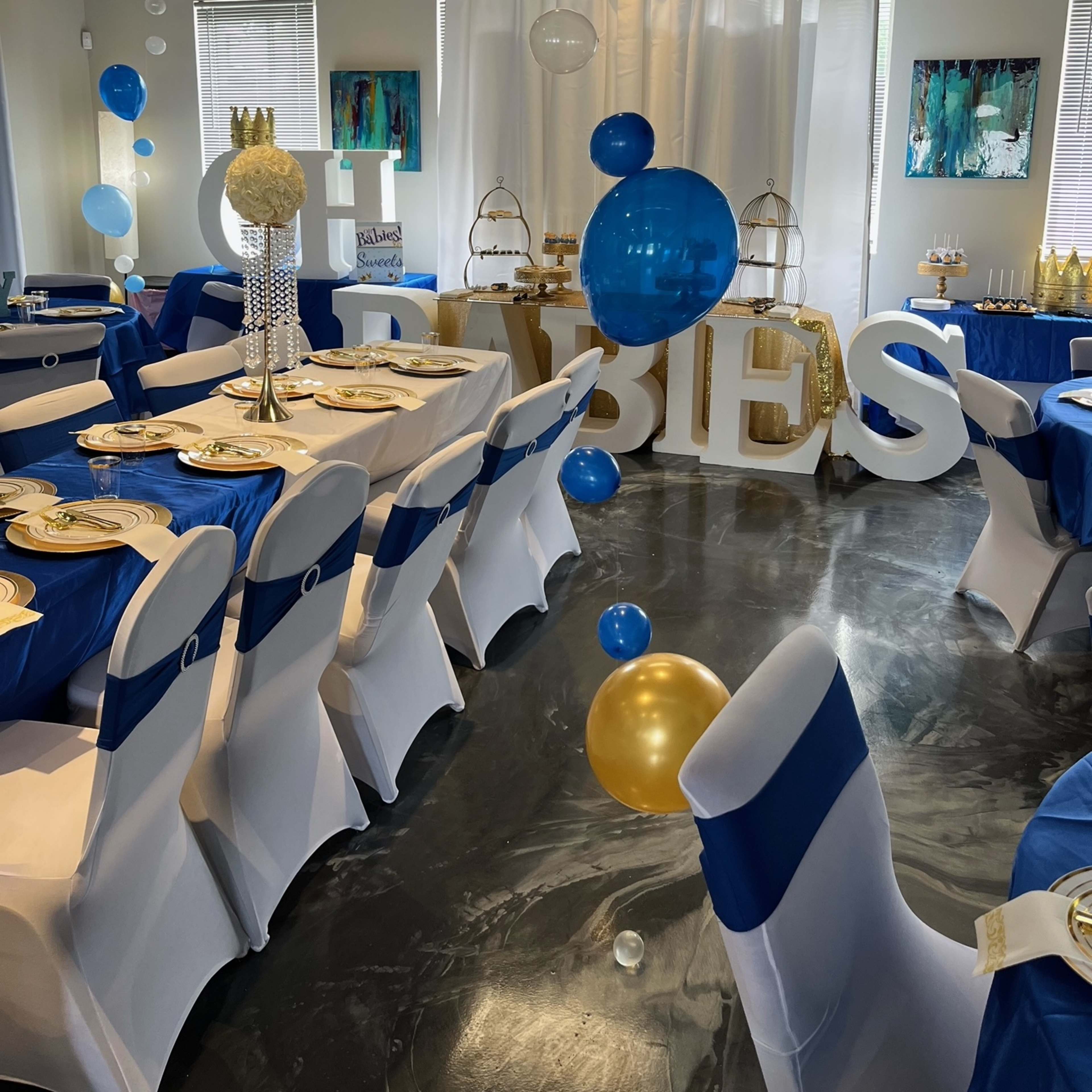 A festive event space decorated with blue and gold balloons, large letters spelling "BABIES," and tables set with blue tablecloths and white chair covers.