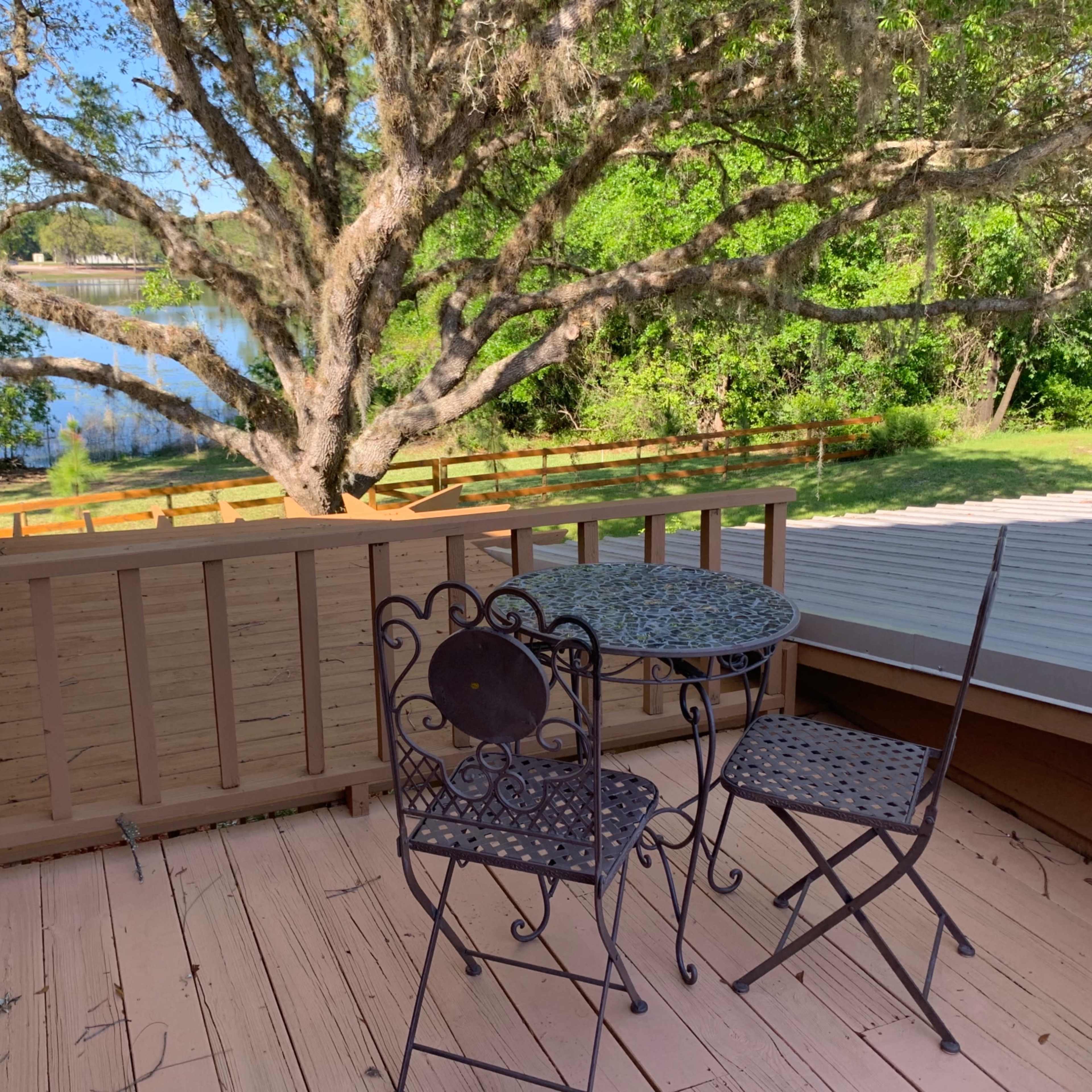 A small balcony features a wrought iron table and chair set overlooking a tranquil body of water and lush greenery.