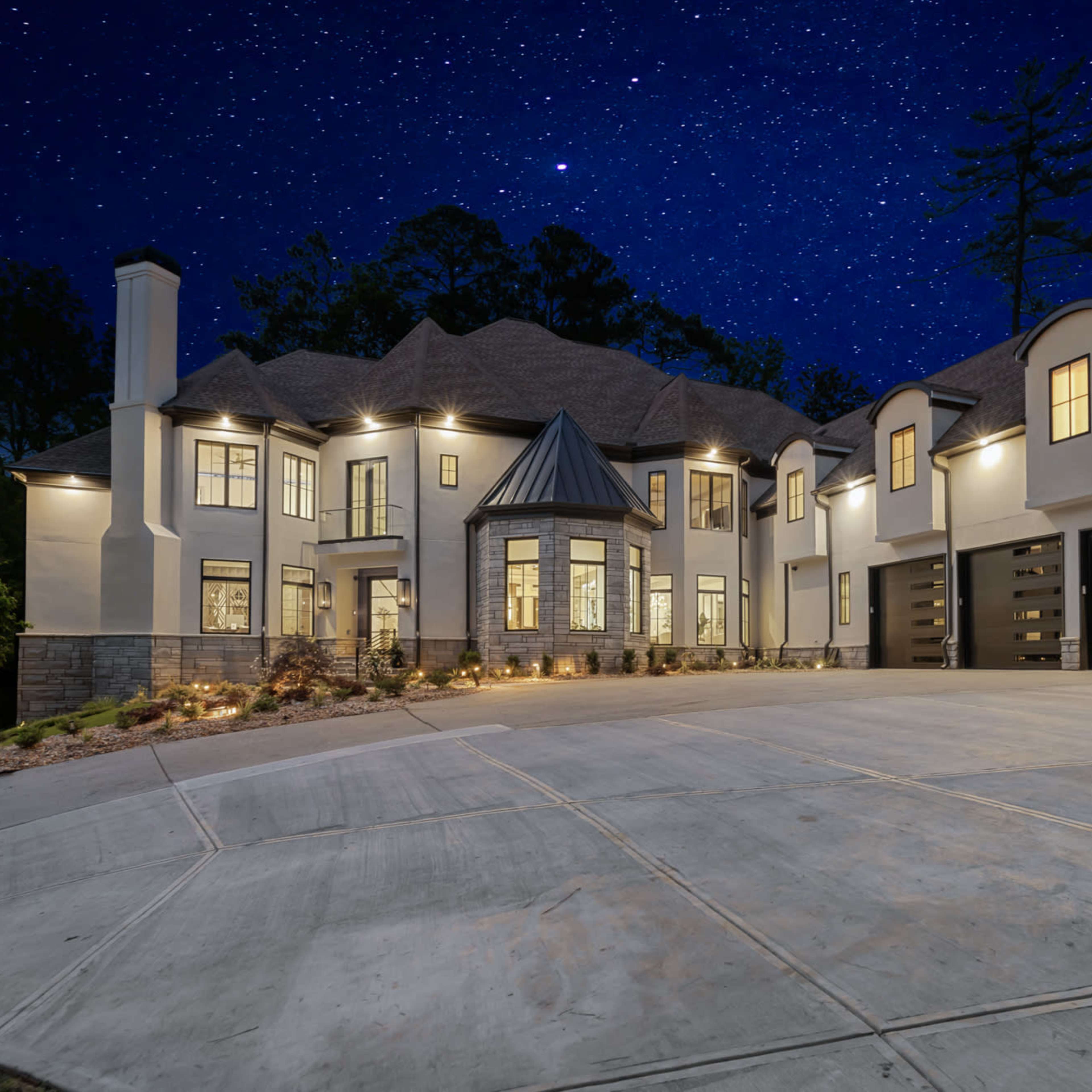 A large, modern house with multiple gables is illuminated at night, showcasing a spacious driveway and starry sky above.