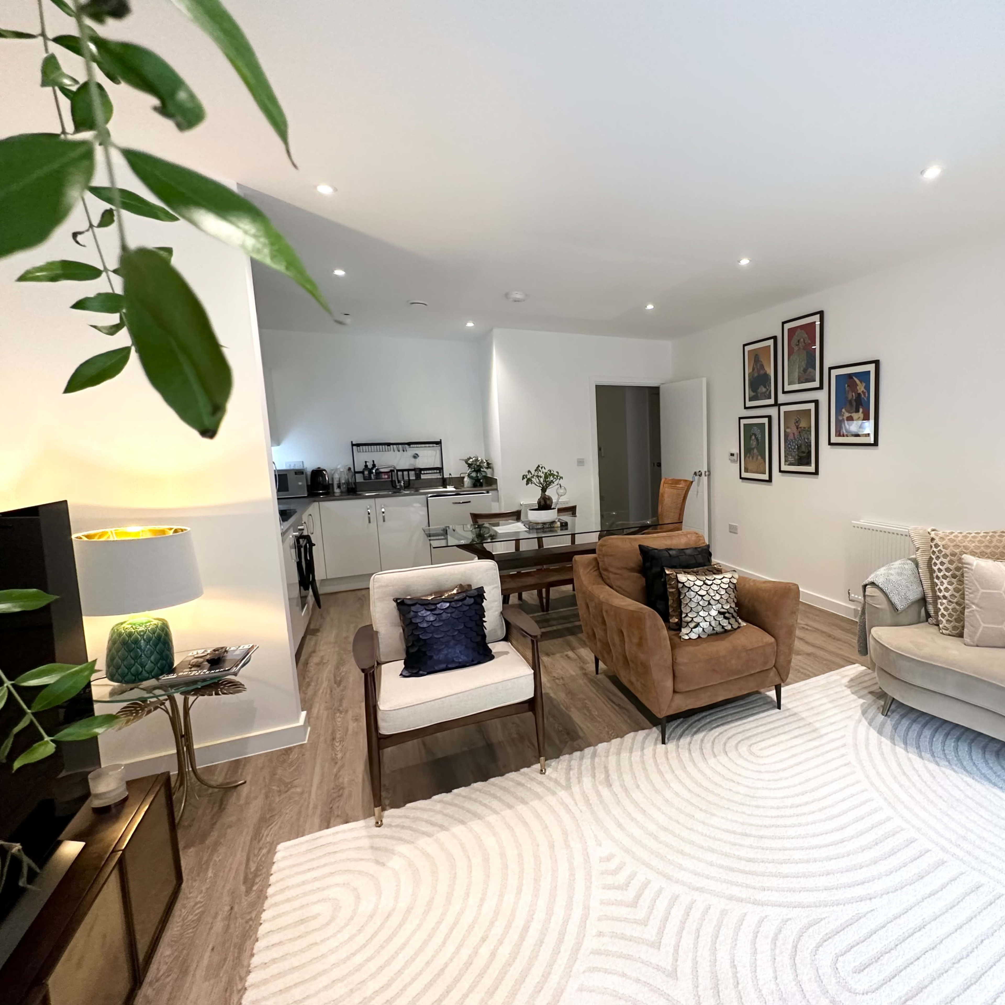The living room features a light-colored sofa, two armchairs, a round rug, and framed artworks on the wall, with a small dining area visible in the background.