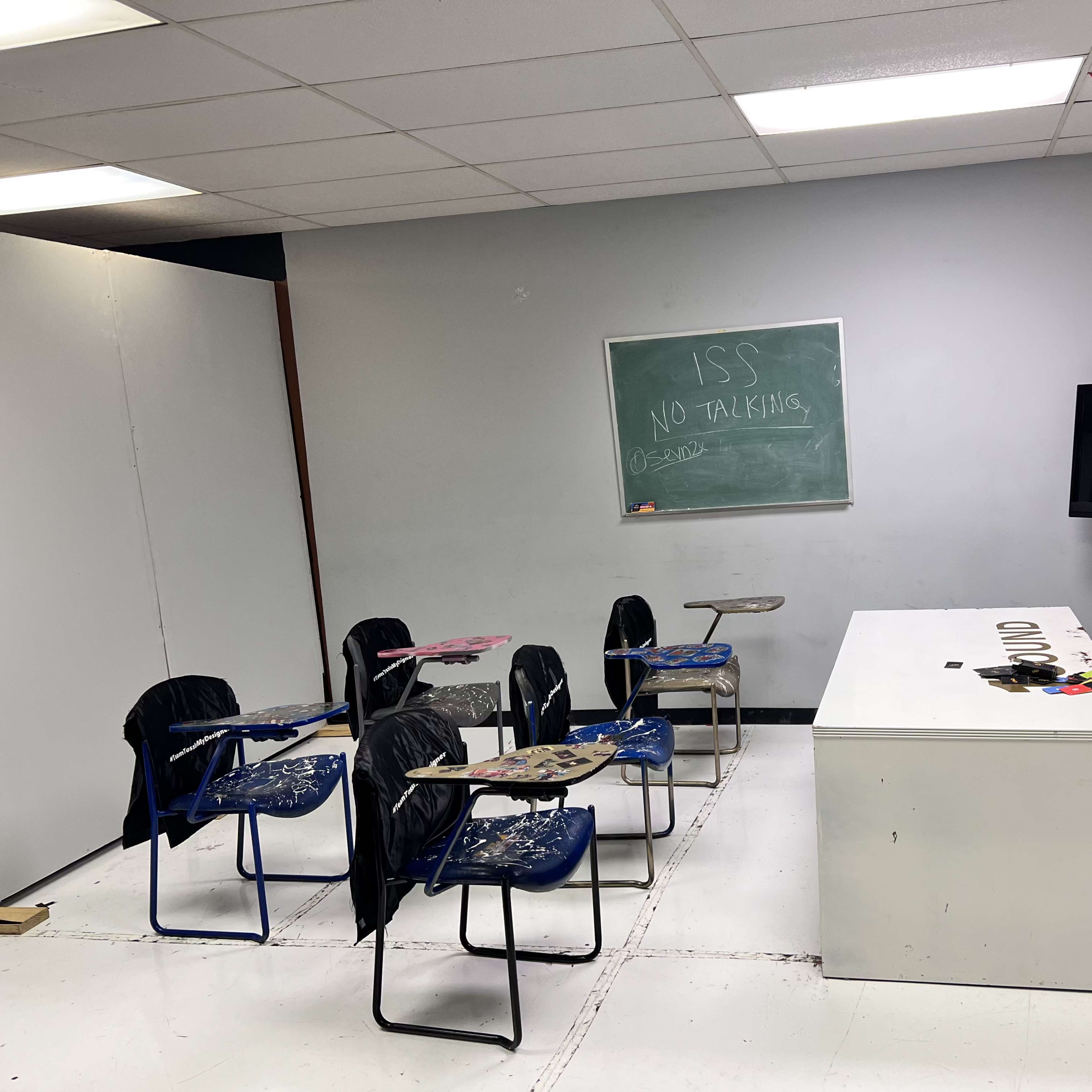 An empty classroom features several desks, a large white desk, a television, and a chalkboard with rules written on it.