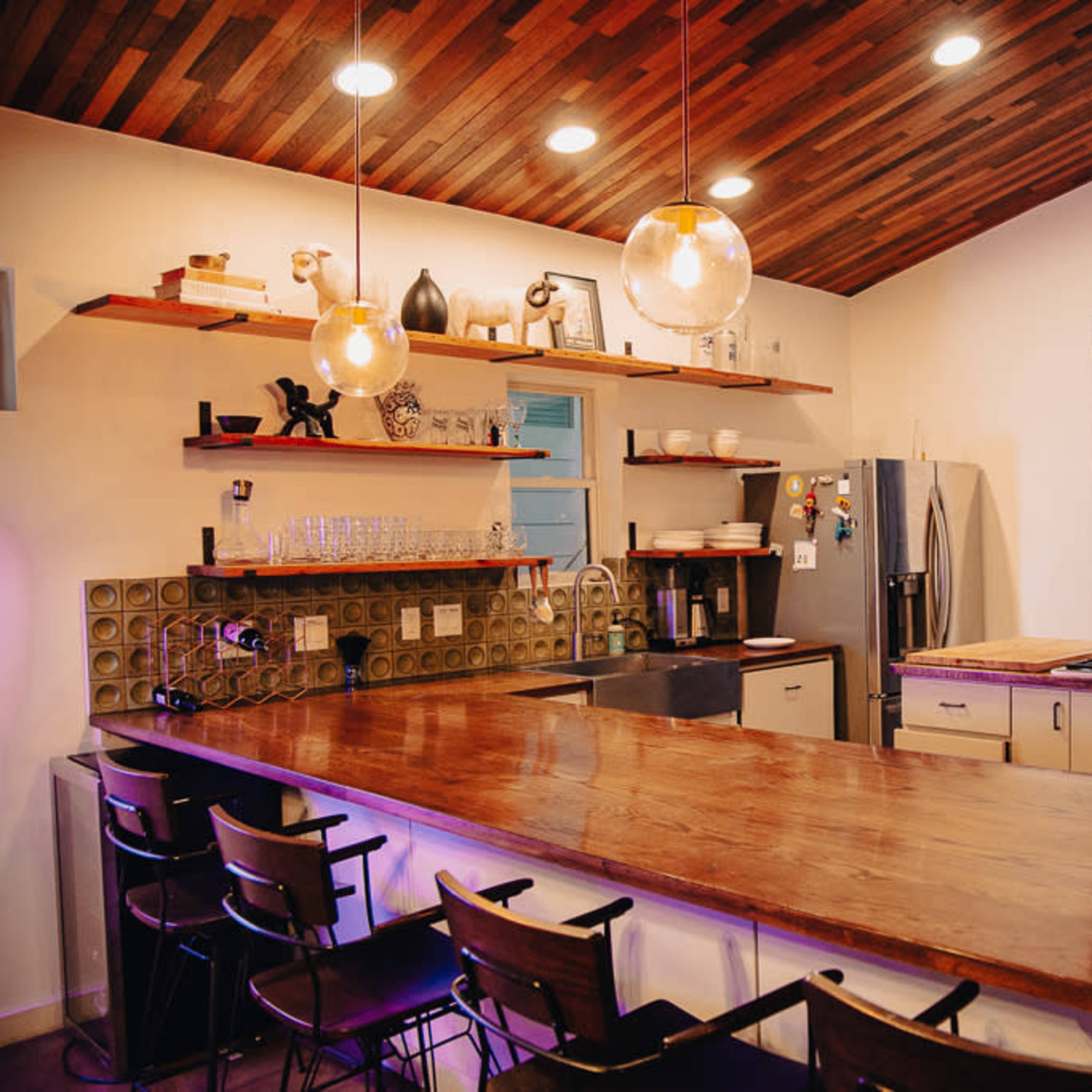 A modern kitchen features a large wooden counter, shelves with kitchenware, and a stainless steel refrigerator, with a view of the outdoors through a wide open window.