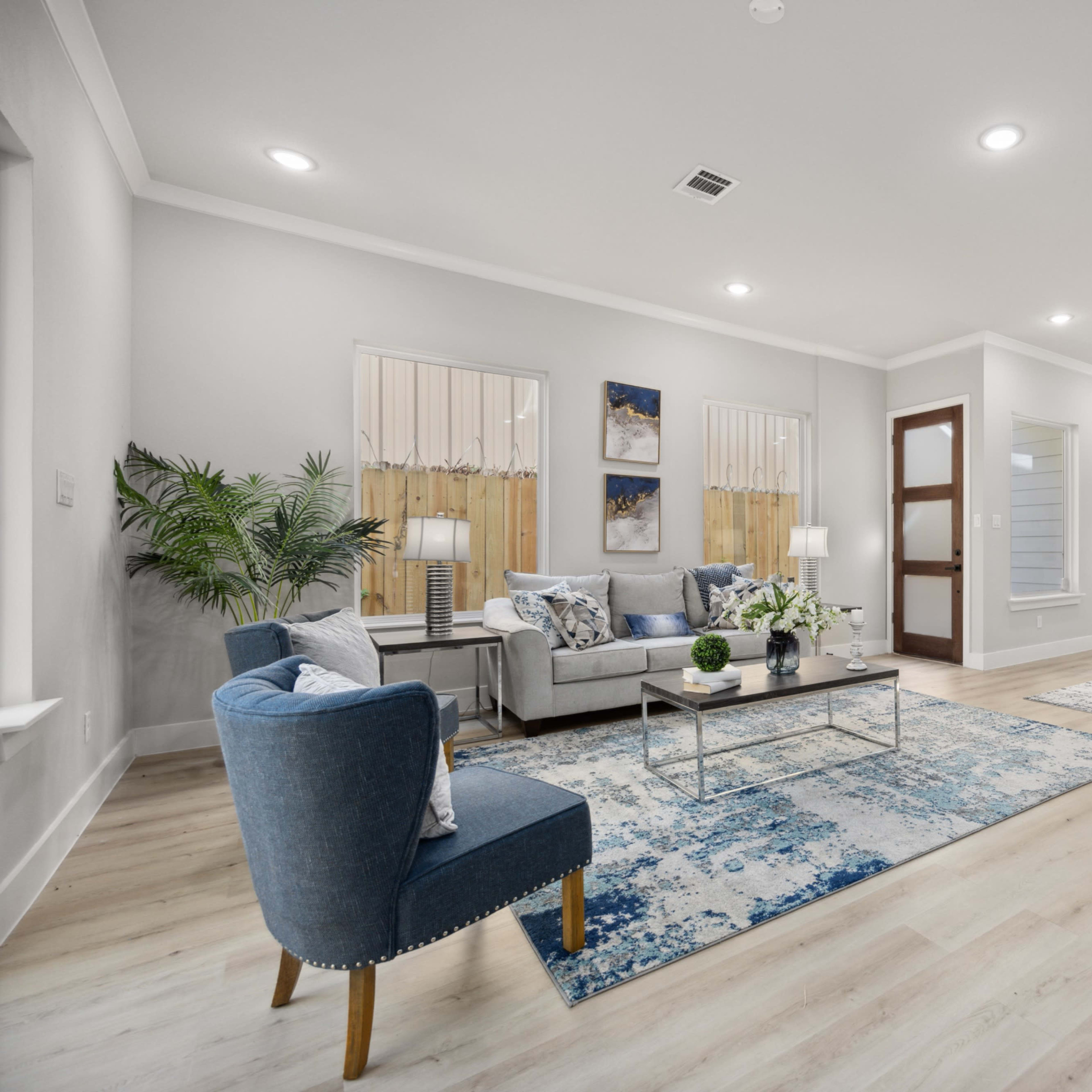 A spacious, modern living area with a blue and white color scheme, featuring a seating arrangement, decorative plants, and an open kitchen that connects to a dining area.