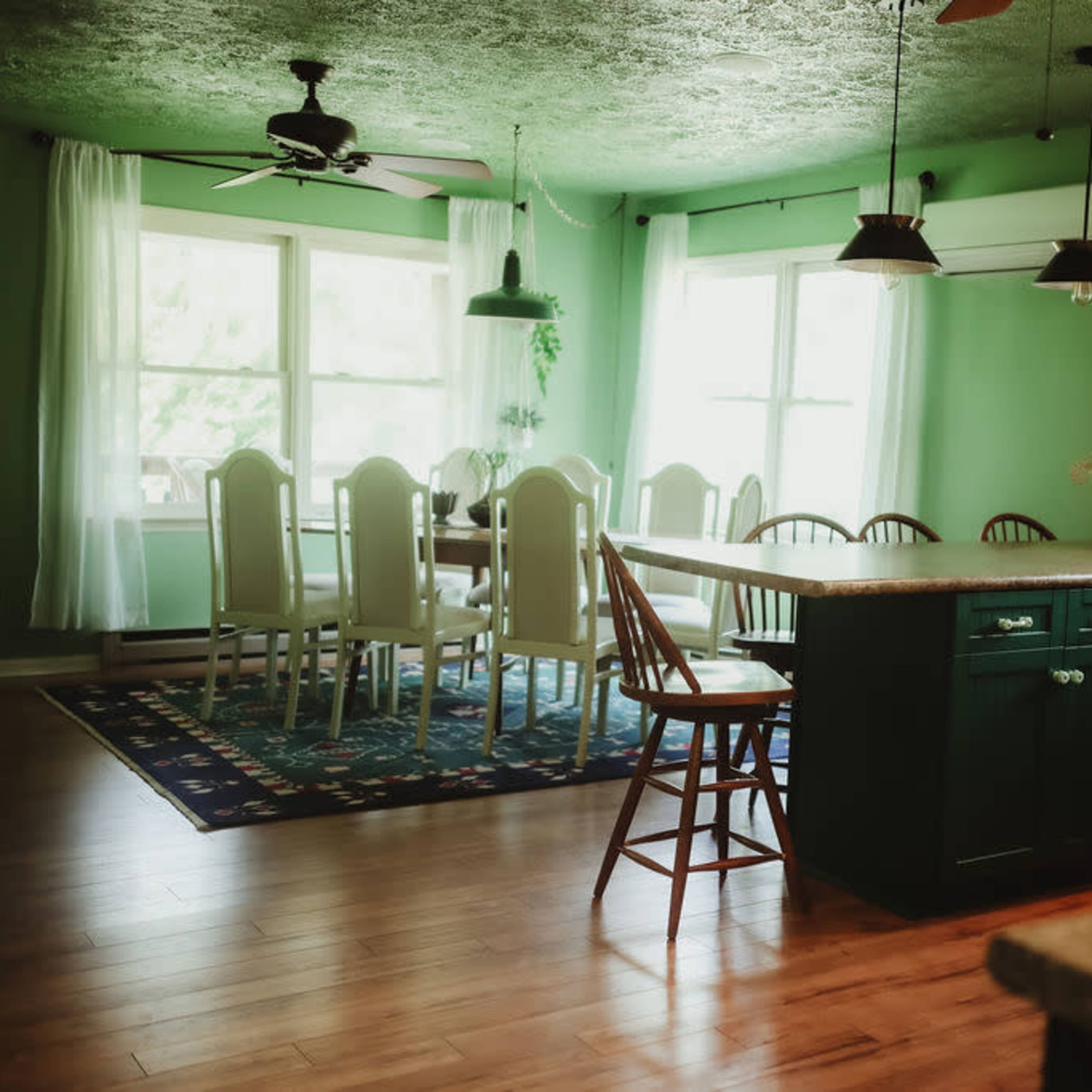 A spacious kitchen features a wooden dining table with chairs, green walls, large windows, and wooden floors.