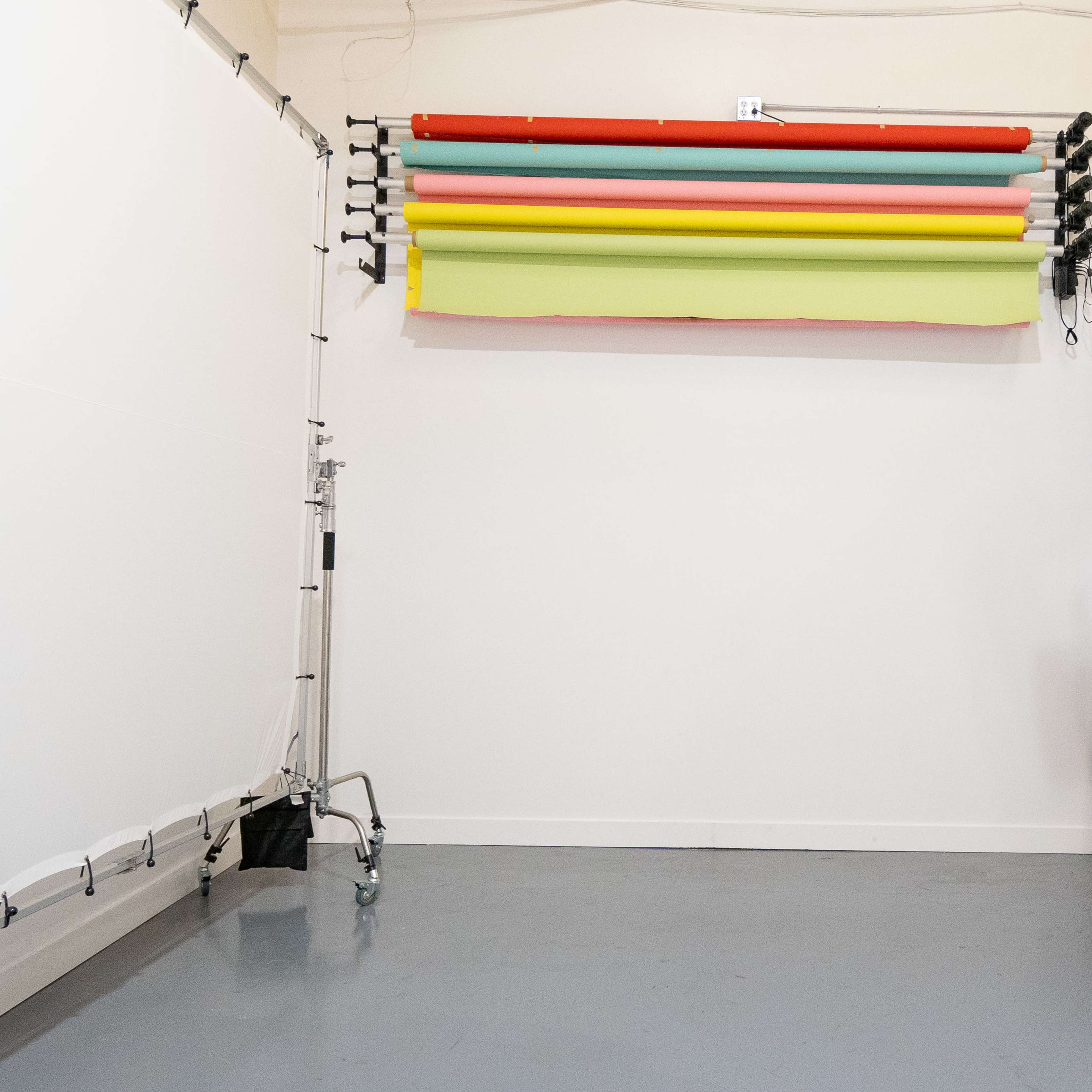A ladder stands next to a wall-mounted backdrop system with various colored rolls in an empty studio space.