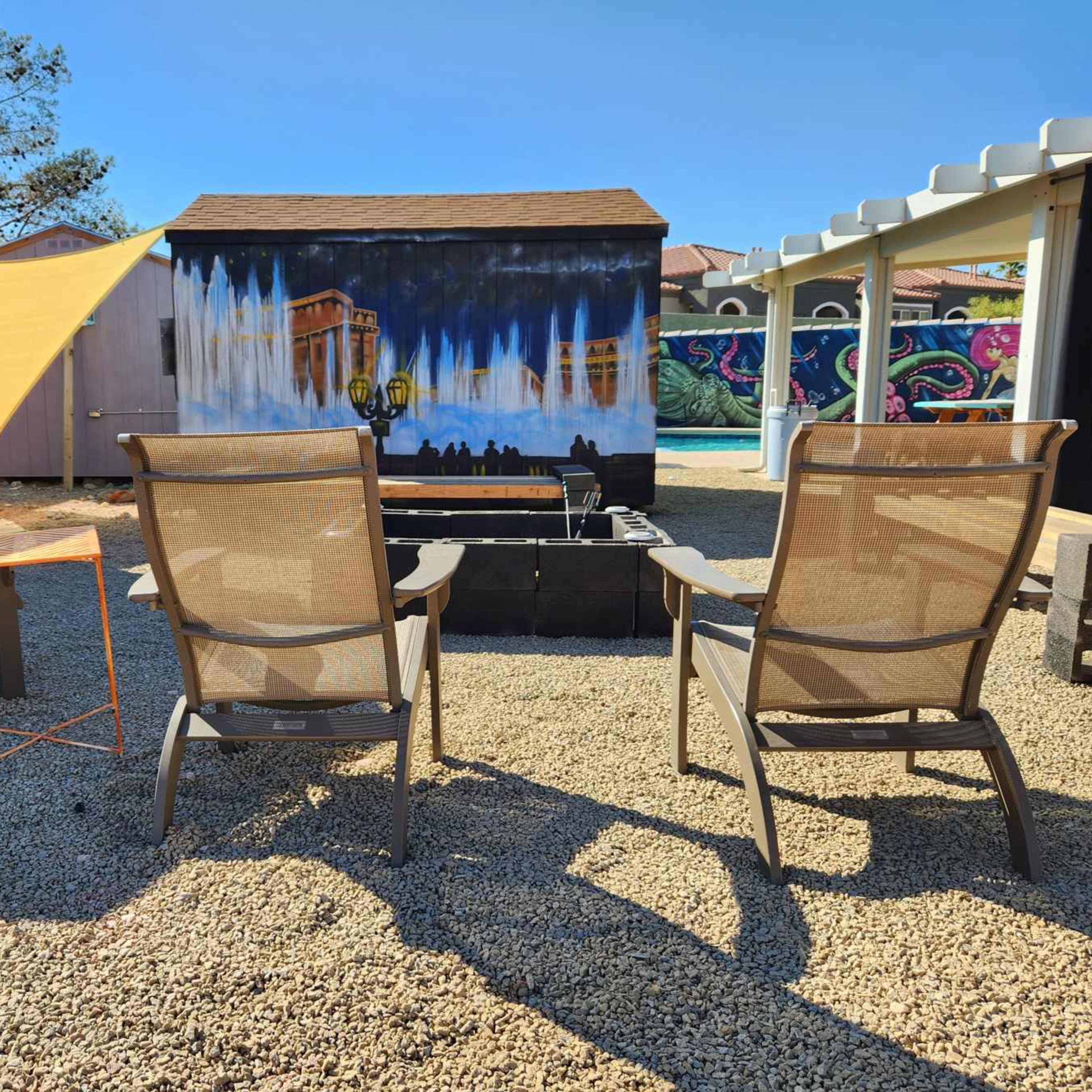 Three lounge chairs are arranged around a small table in a gravel courtyard beside colorful murals on nearby walls.