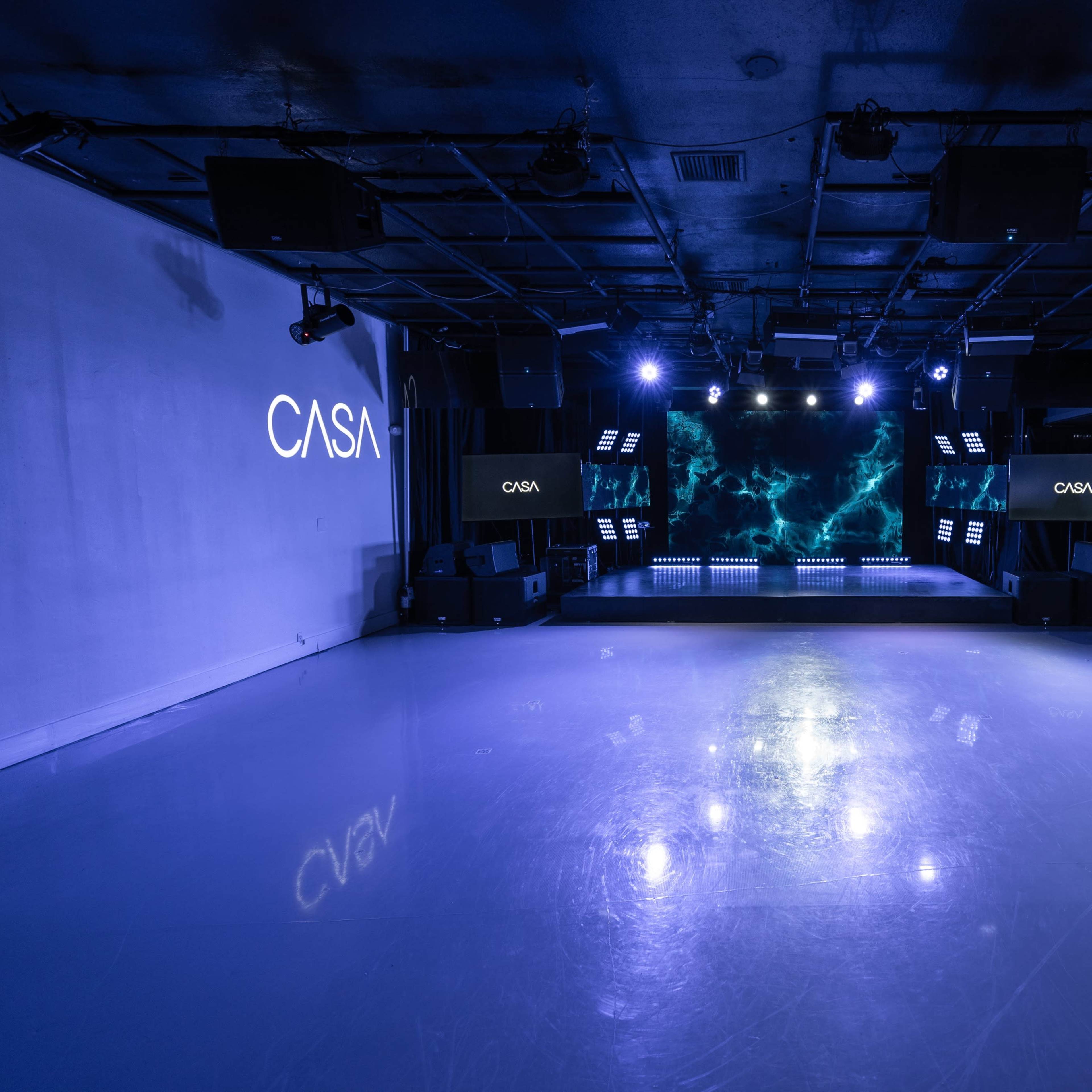 A modern event space with a shiny reflective floor, lit by blue lights and featuring a stage at the front.