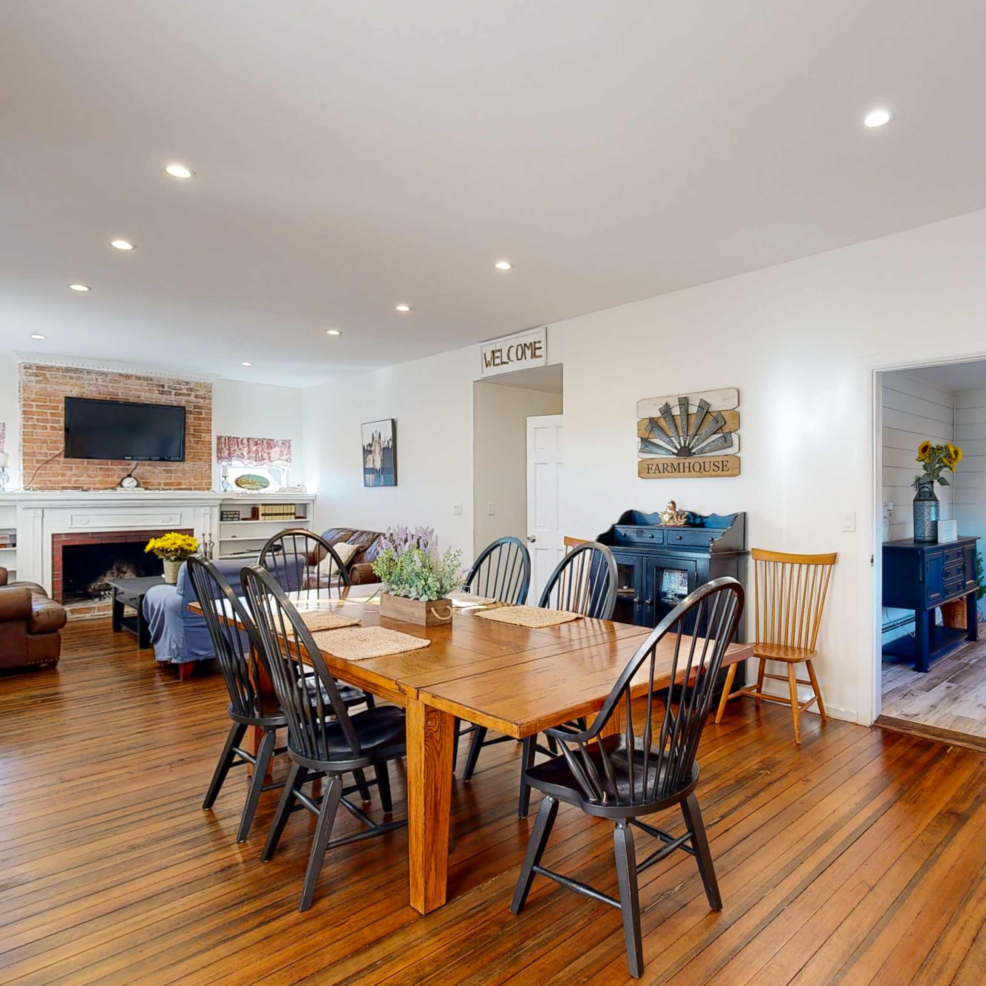 A spacious dining area with a wooden table surrounded by black chairs, a brick fireplace in the background, and a doorway leading to another room.