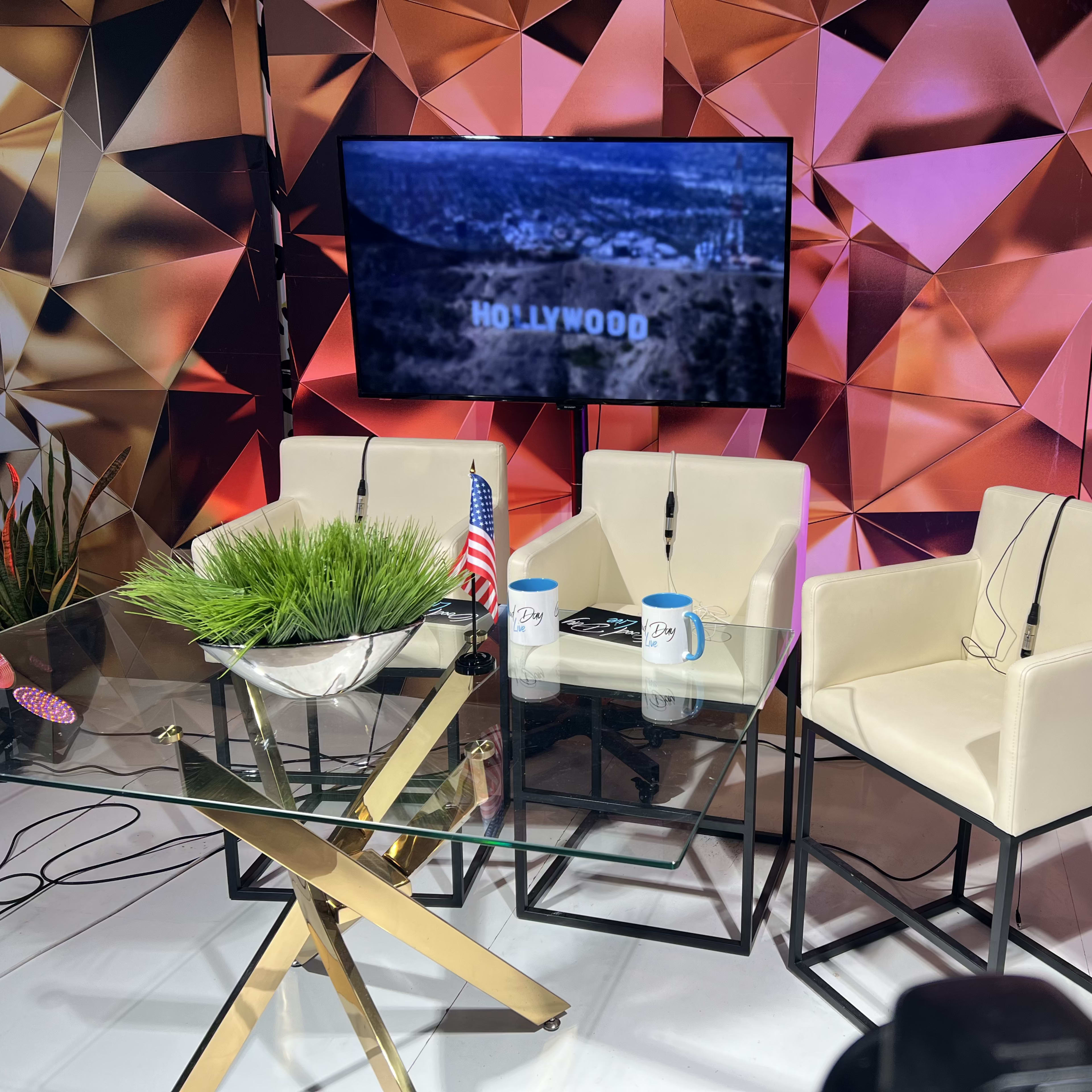 A television interview setup with three white chairs, a glass table, and a screen displaying "HOLLYWOOD" against a geometric backdrop.