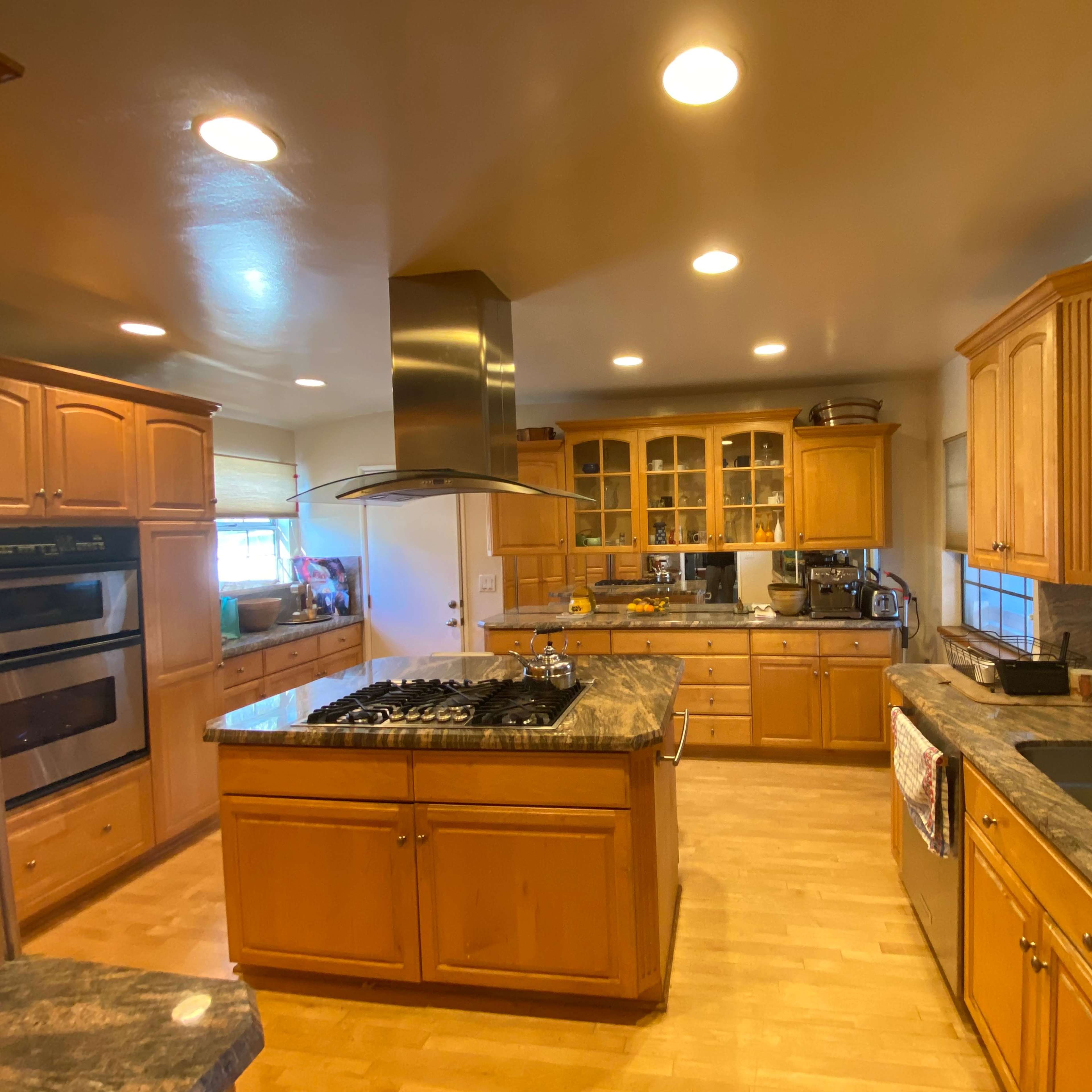 A spacious kitchen features wooden cabinetry, a large island with a gas cooktop, and ample counter space next to double sinks.