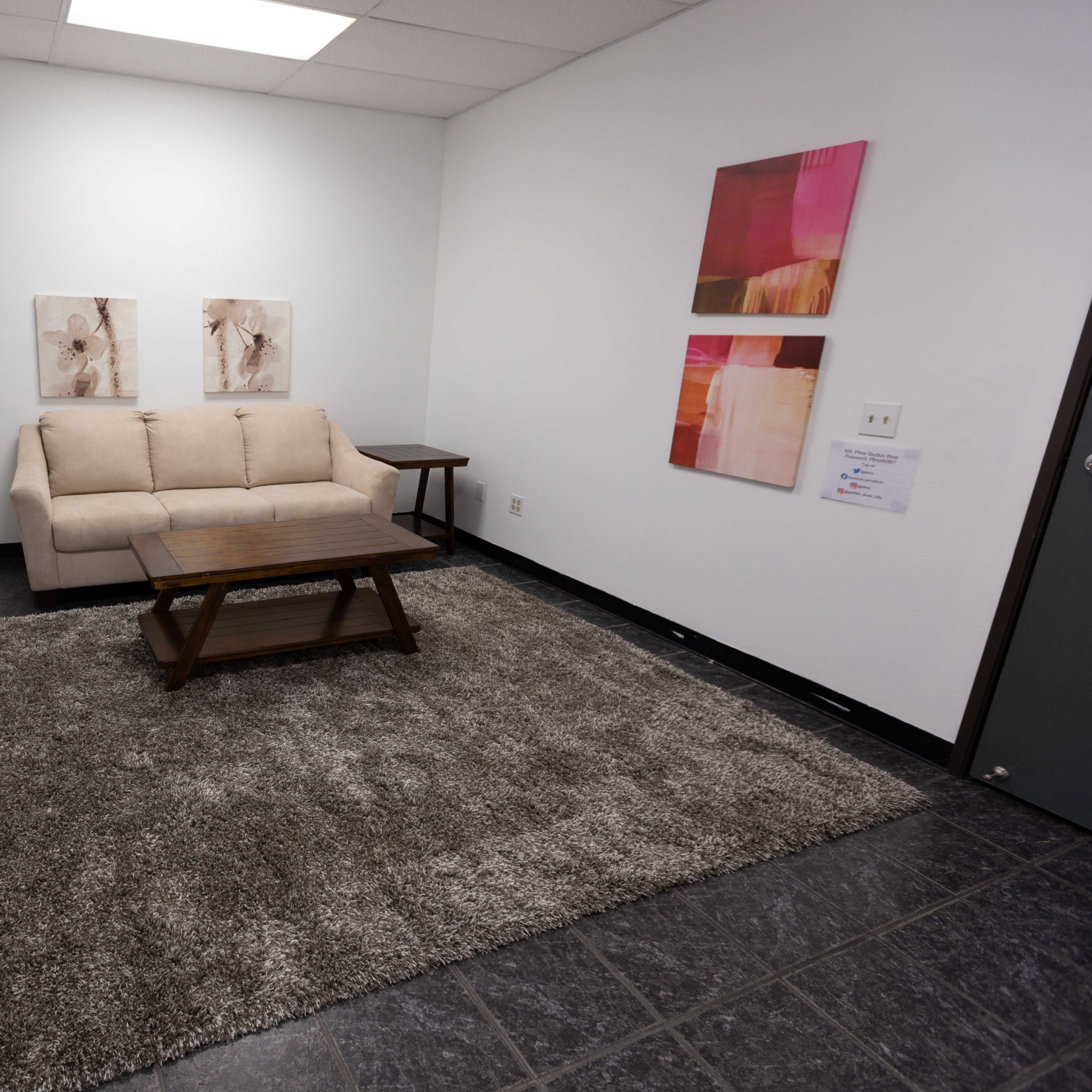 A room with a beige couch, a wooden coffee table, and two pieces of framed artwork on the wall, alongside a grey rug on a tiled floor.