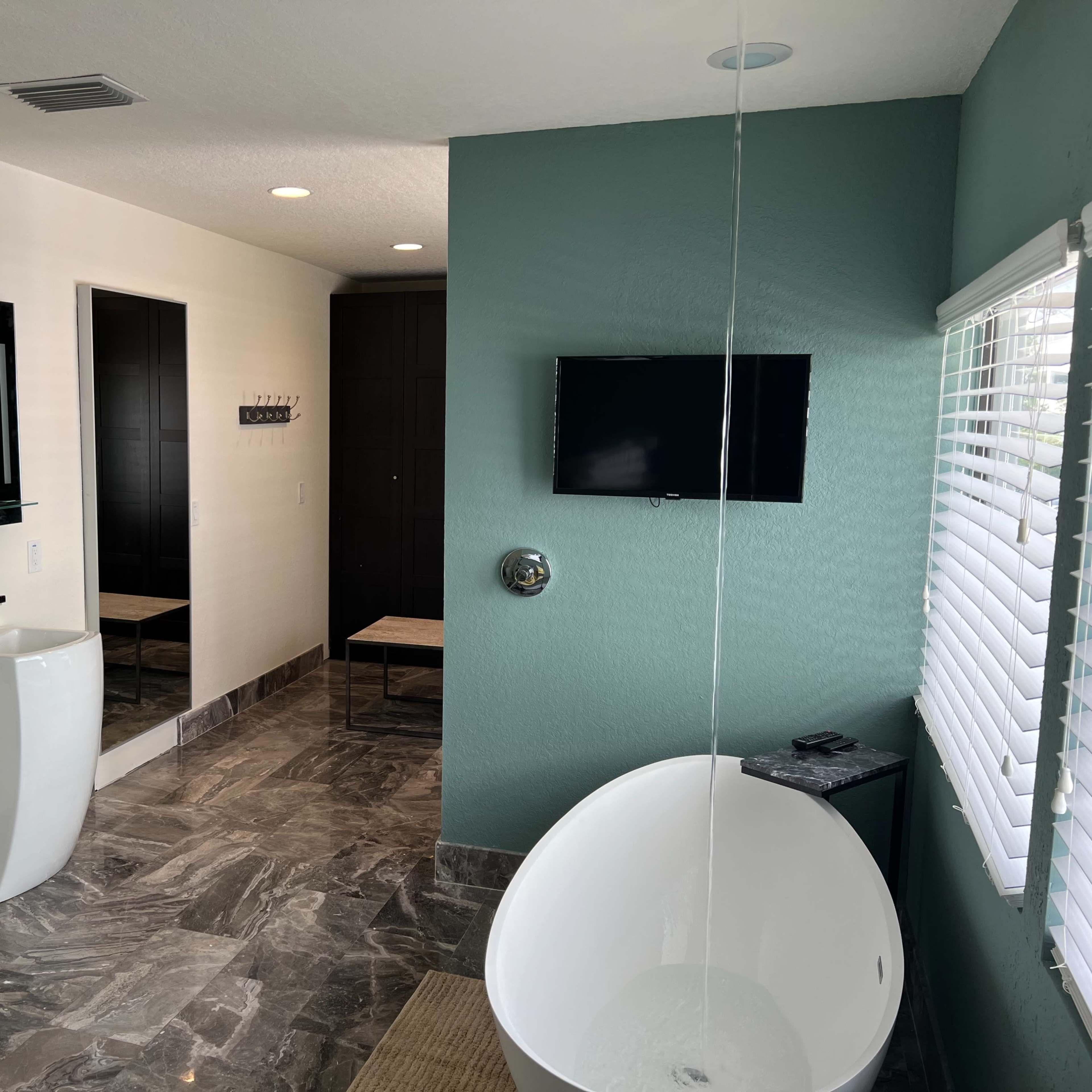 A modern bathroom featuring two white pedestal sinks, a freestanding bathtub, a wall-mounted television, and slate-colored walls.