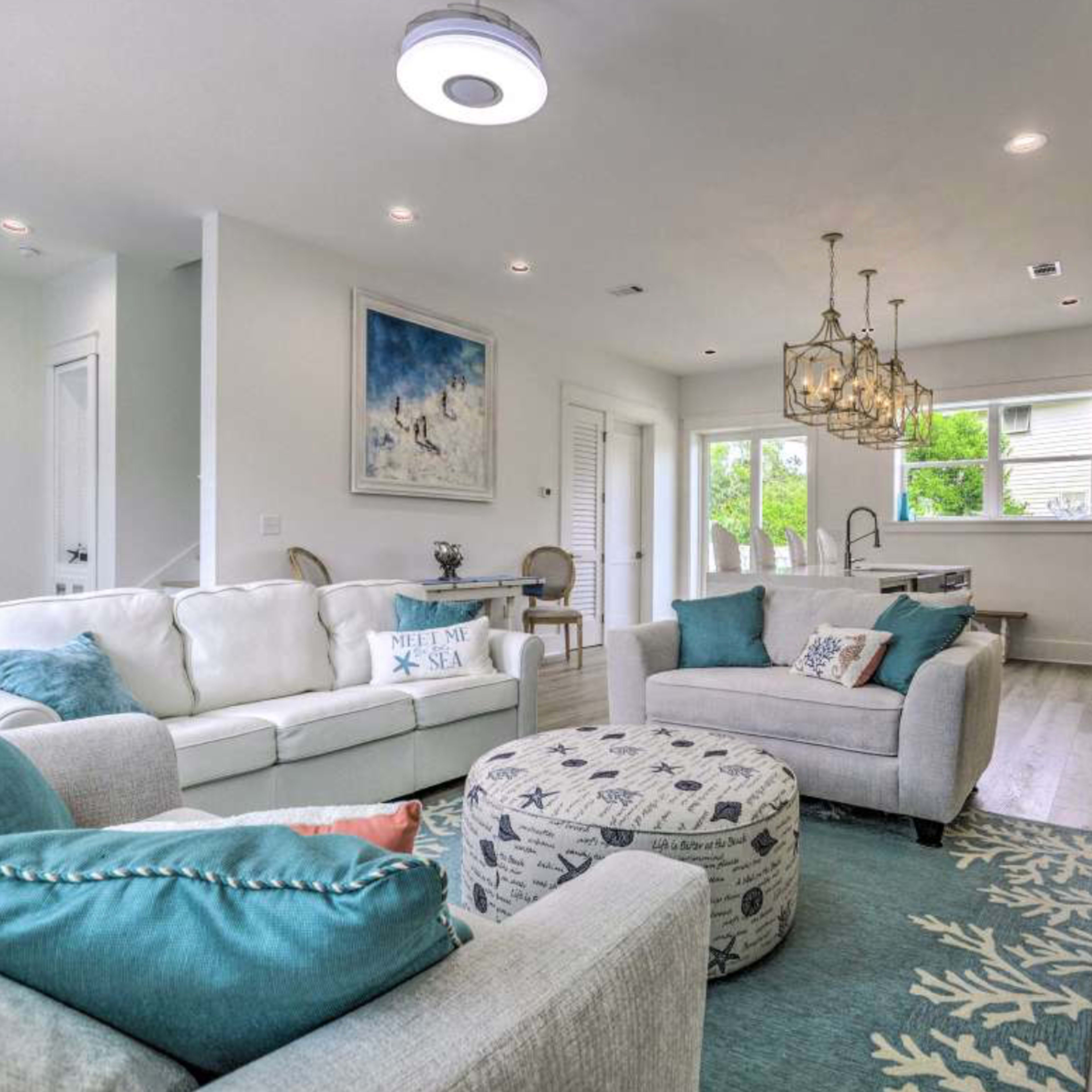 A bright and modern living room featuring a white sofa, a patterned area rug, a circular ottoman, and a stylish dining area with a chandelier.