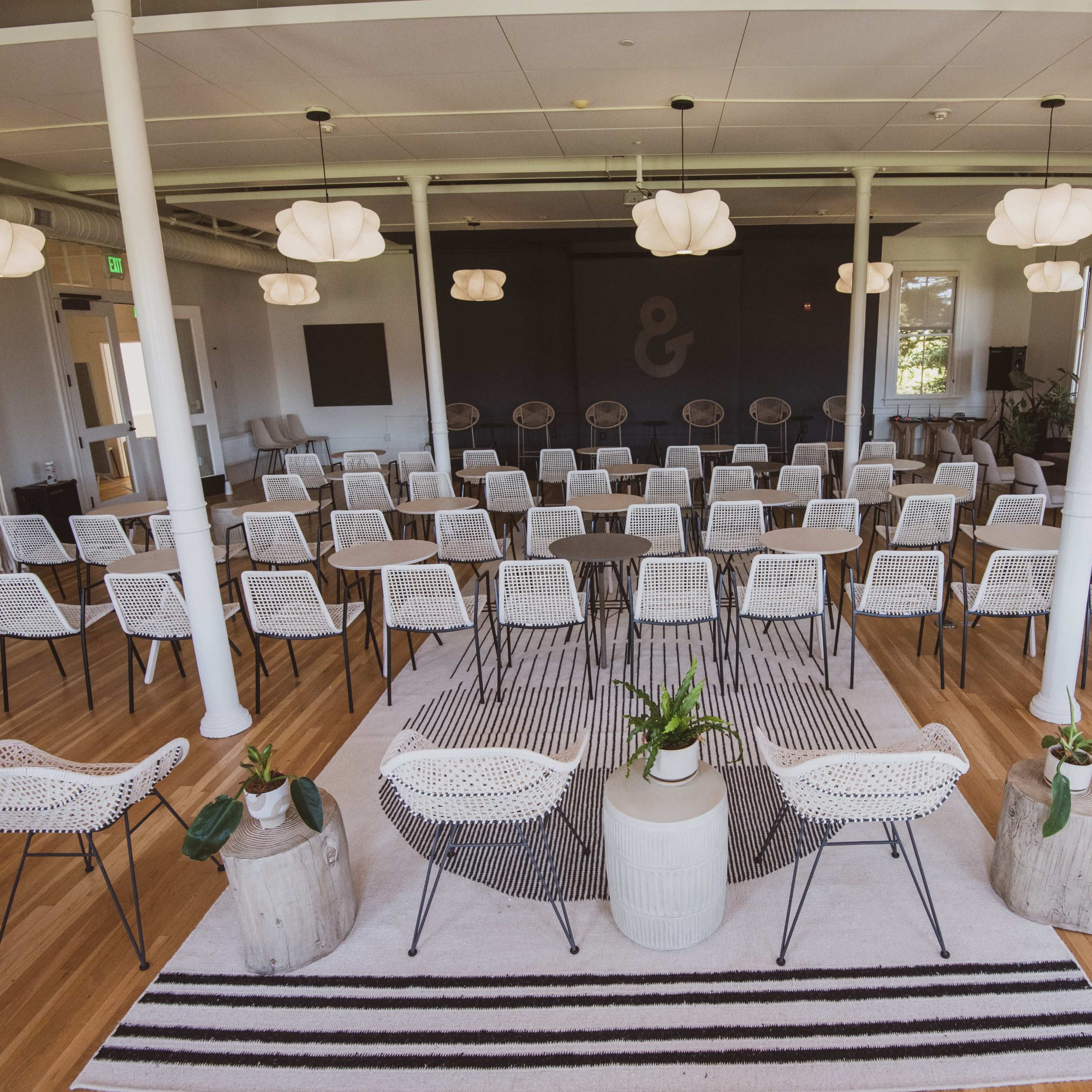 A modern event space is arranged with rows of chairs facing a central stage, surrounded by decorative elements including a rug and plants.
