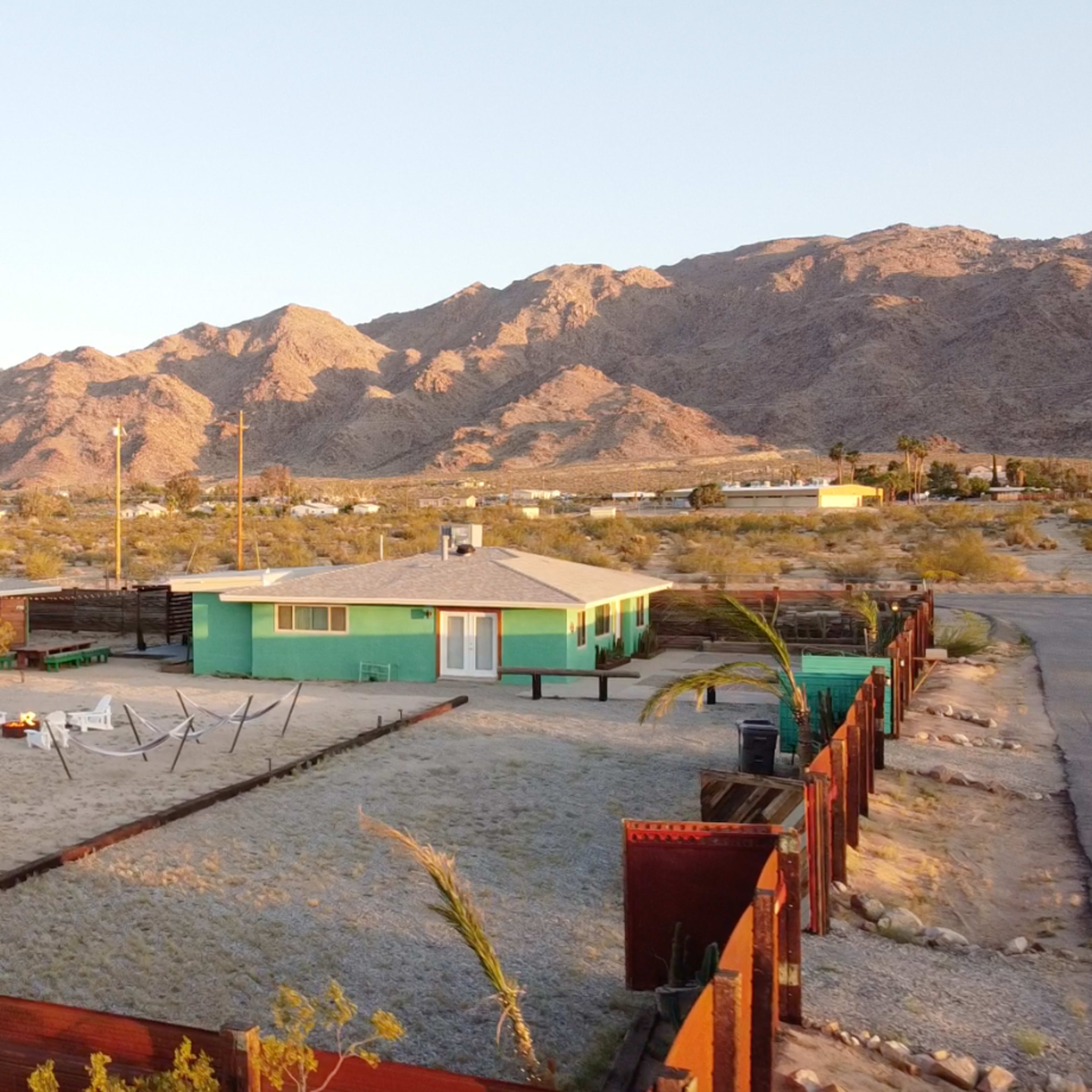 A green house with a sandy yard and white lounge chairs is surrounded by a wooden fence, with mountains in the background and a clear sky.