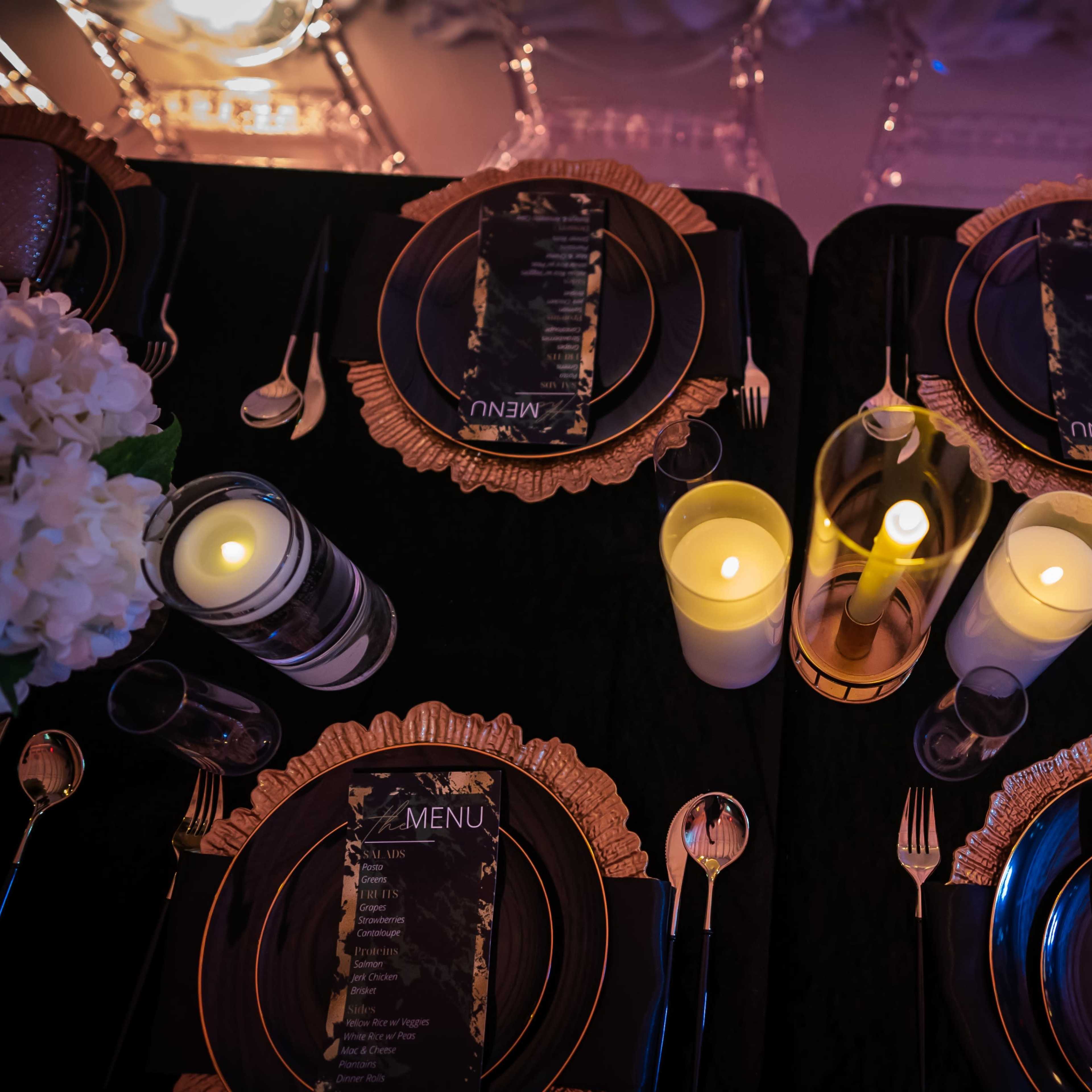 An elegantly set dining table with black plates, golden cutlery, and lit candles arranged around a central menu card.