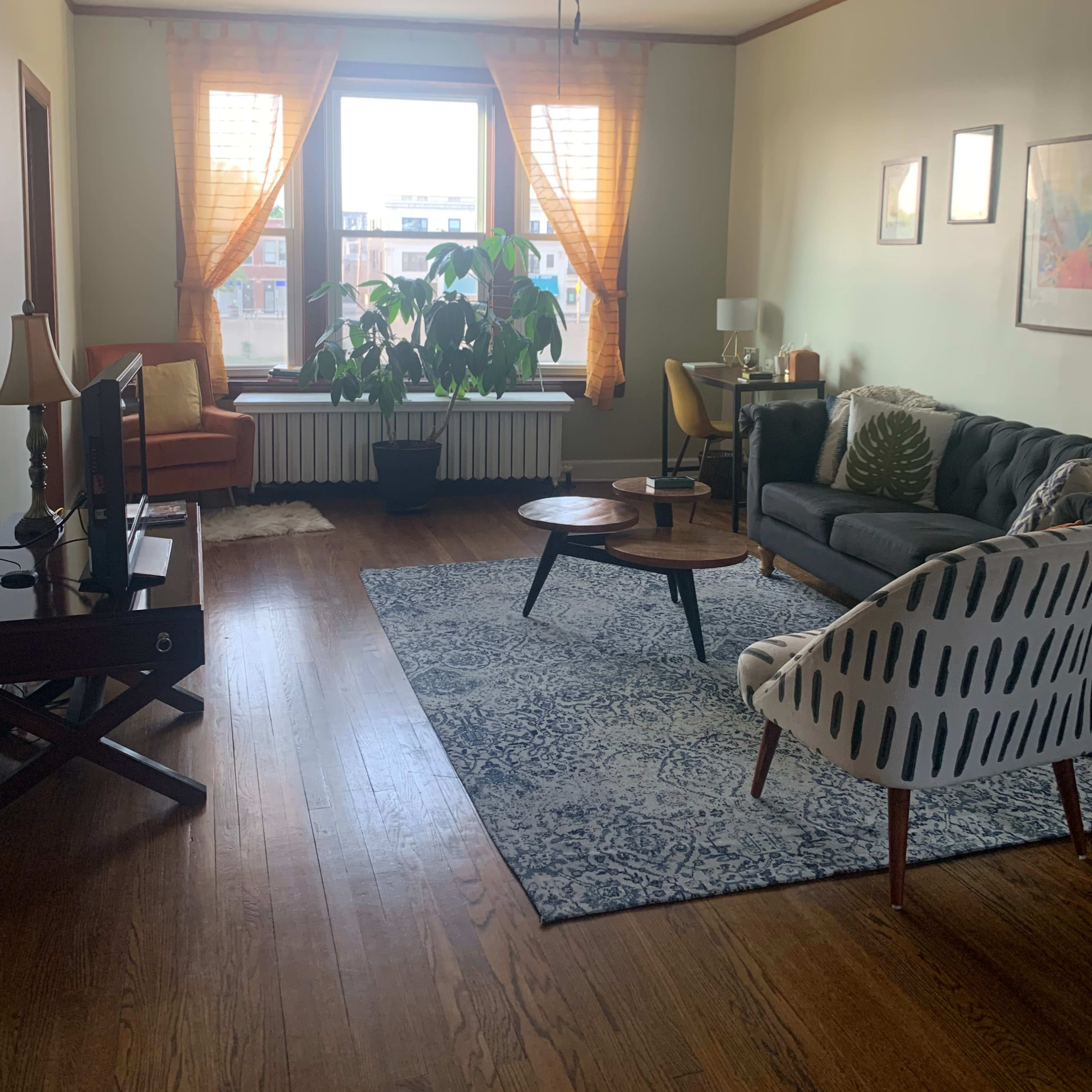 The living room features a large window with sheer curtains, a mix of seating options, a coffee table, and a patterned area rug on hardwood flooring.