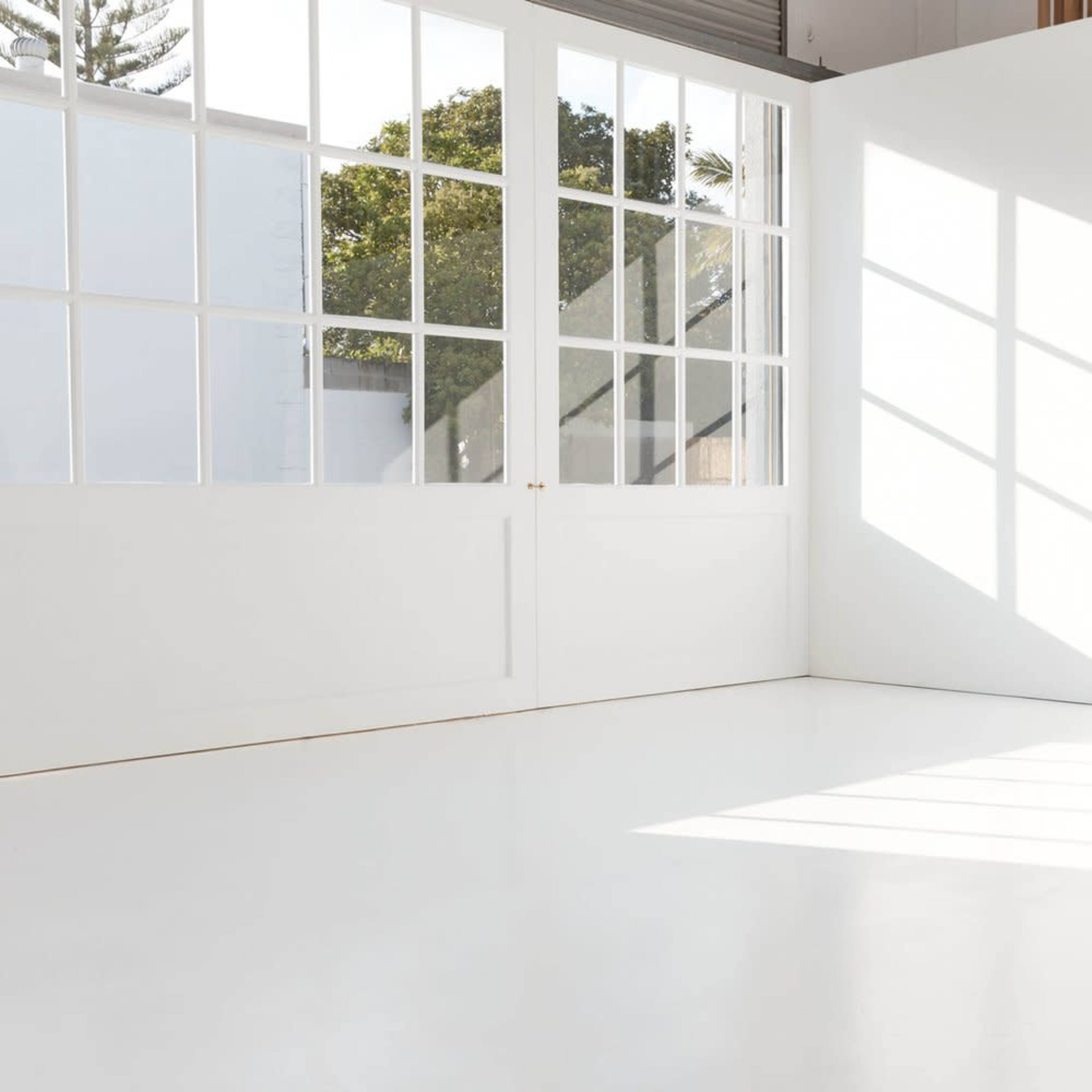 A bright, empty room features large windows with sunlight casting shadows on the white floor.