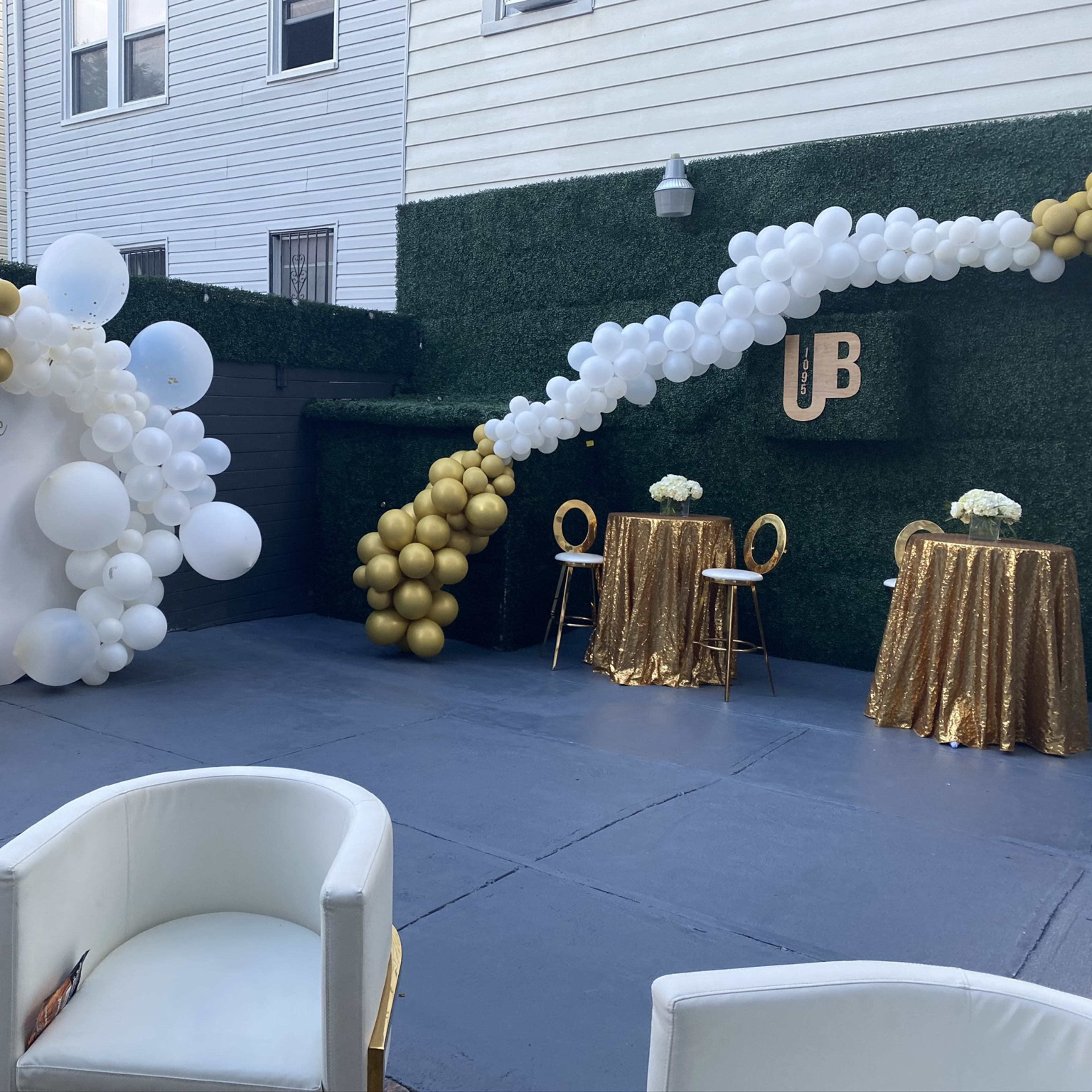 A decorated outdoor space featuring a gold and white balloon arch, a circular backdrop with the name "Michelle," a golden throne chair, and two tables with gold sequined tablecloths.