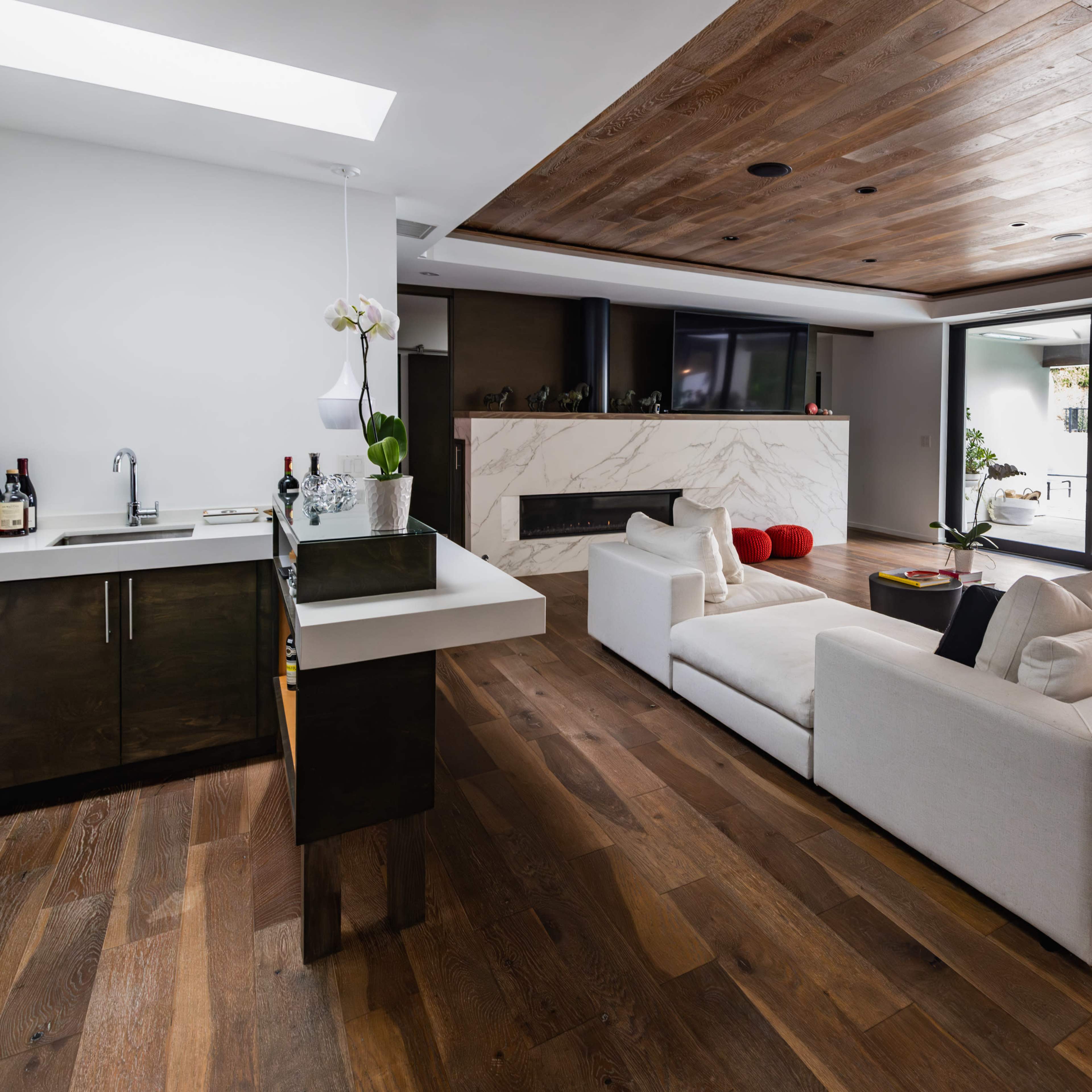 A modern living room features a white sectional sofa, a marble fireplace, a bar area with various bottles, and large glass doors leading to an outdoor patio.