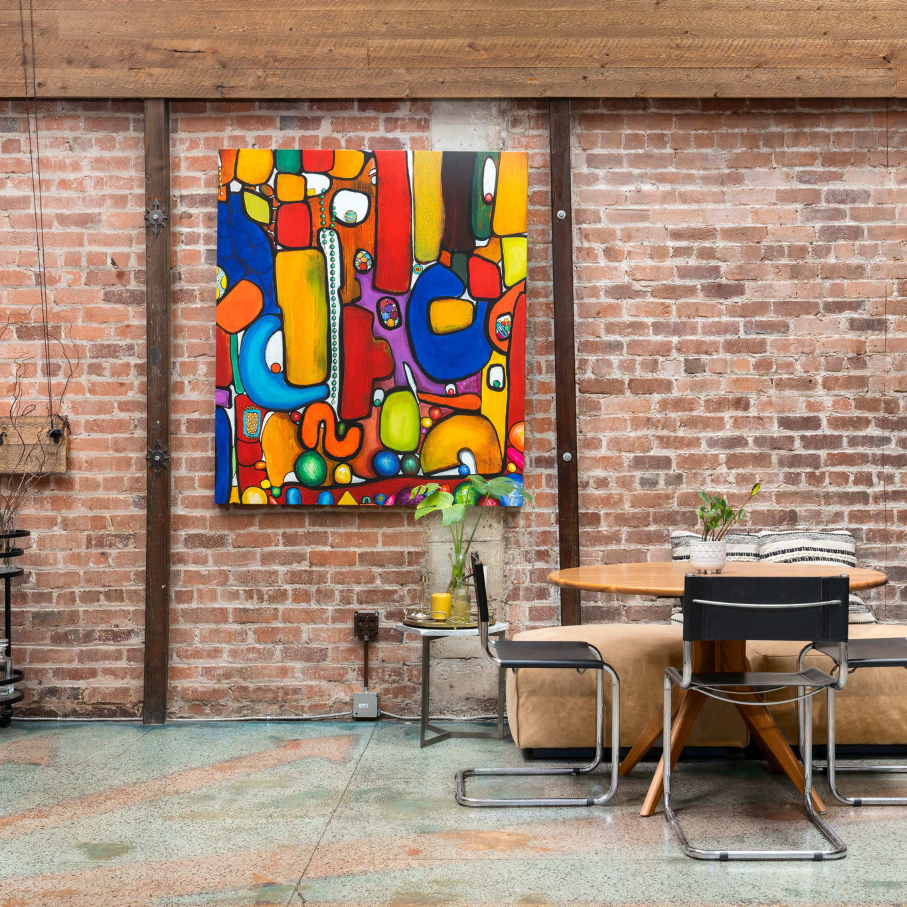 A round wooden dining table with black chairs is positioned against a brick wall featuring a colorful abstract painting.