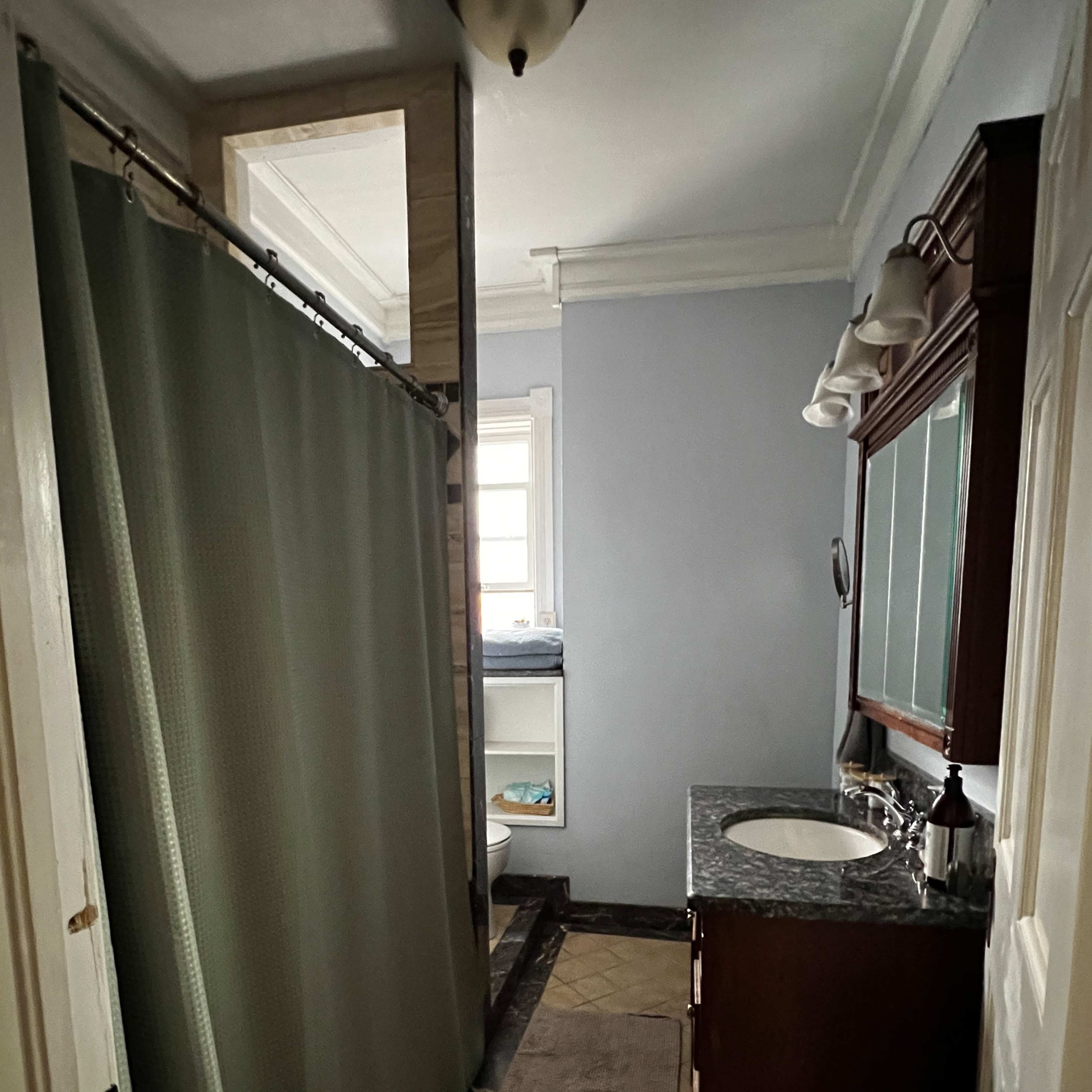 A bathroom featuring a shower curtain, a sink with a mirror above it, and a toilet positioned near a window.