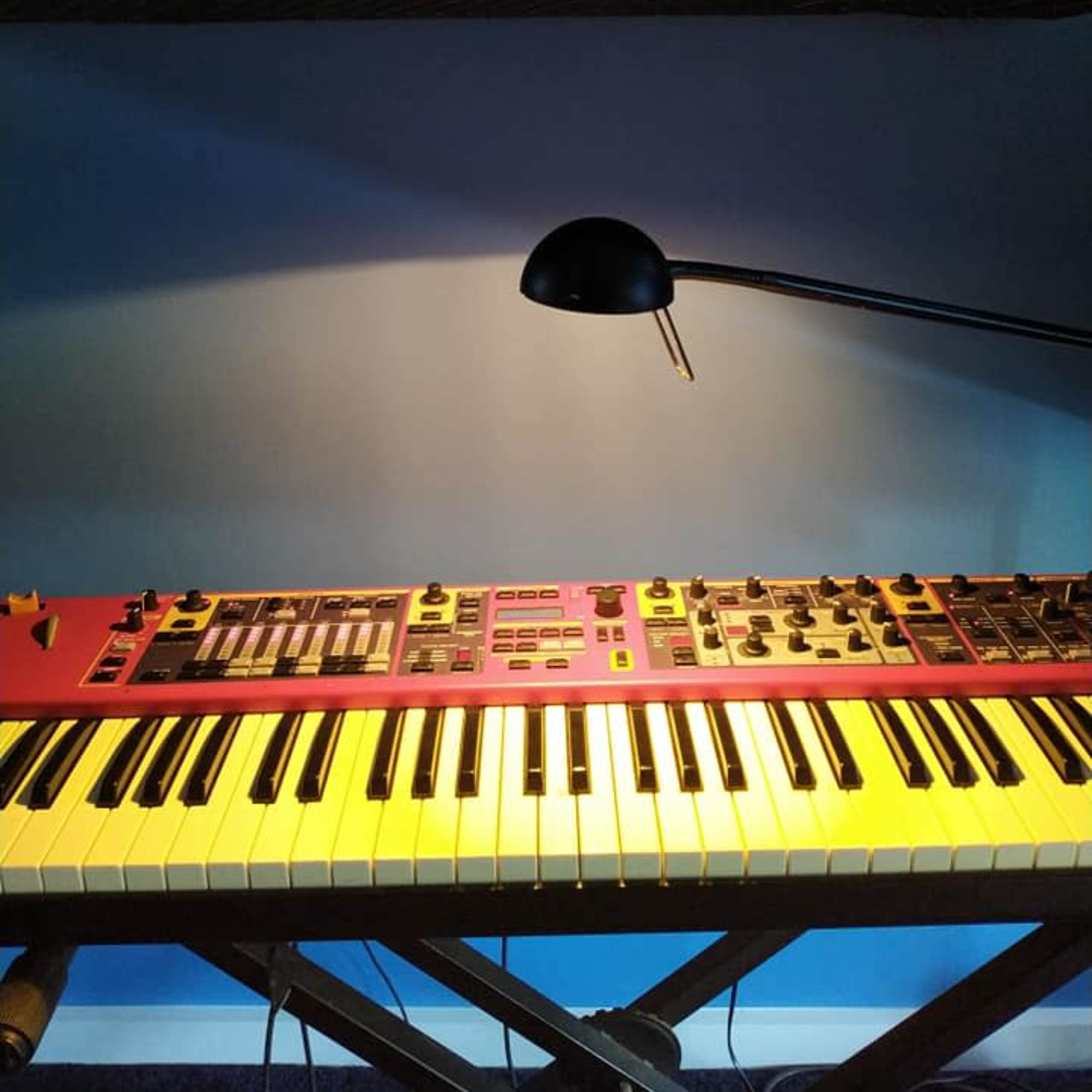 A synthesizer with a red panel and black keys sits on a wooden stand beneath a lamp, against a blue wall.