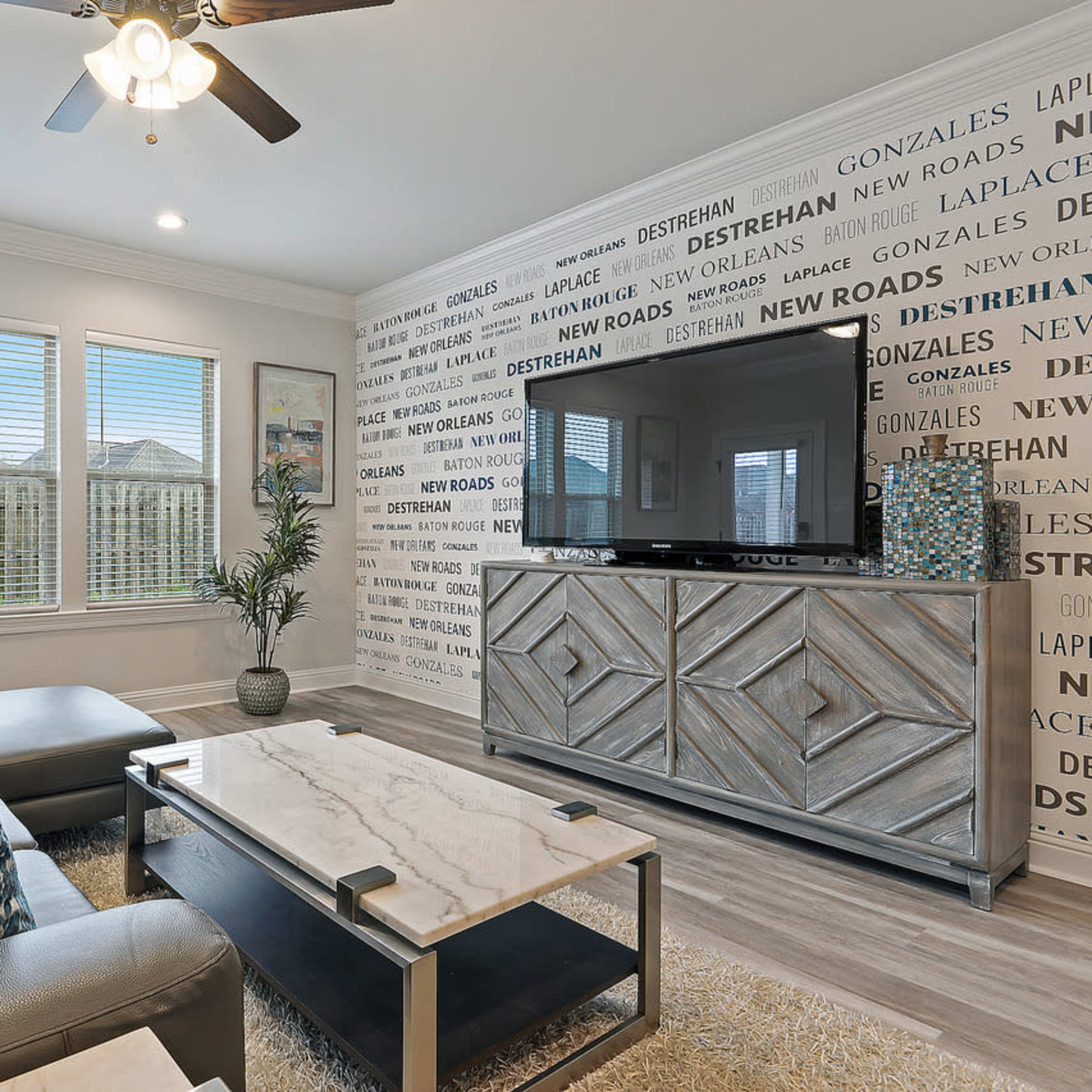 A contemporary living room with a sectional sofa, a decorative coffee table, a large television, and a wall featuring text patterns, complemented by windows overlooking a fenced yard.
