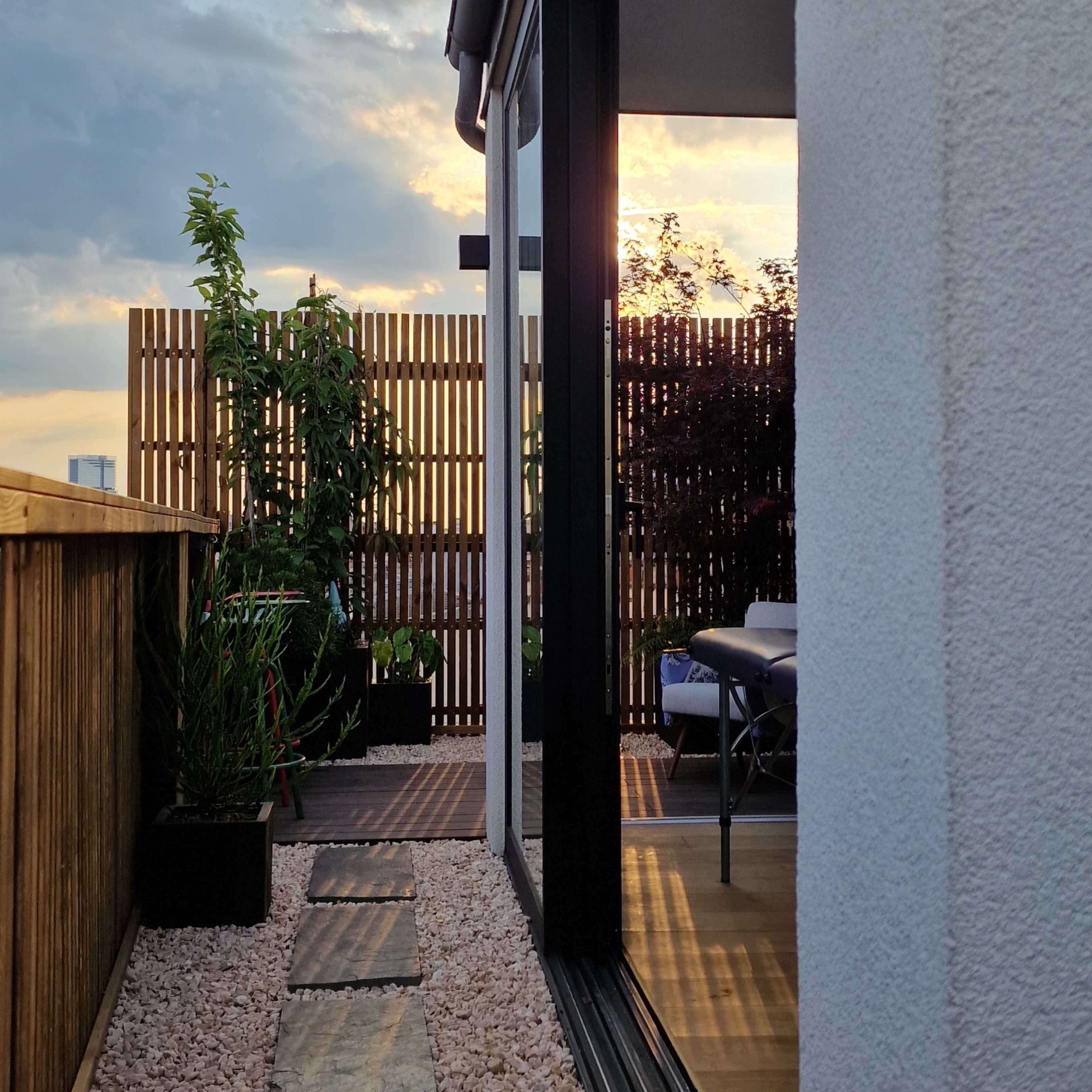 A narrow path lined with stones leading to a modern home with large windows, bordered by plants and a wooden fence, under a cloudy sky at sunset.