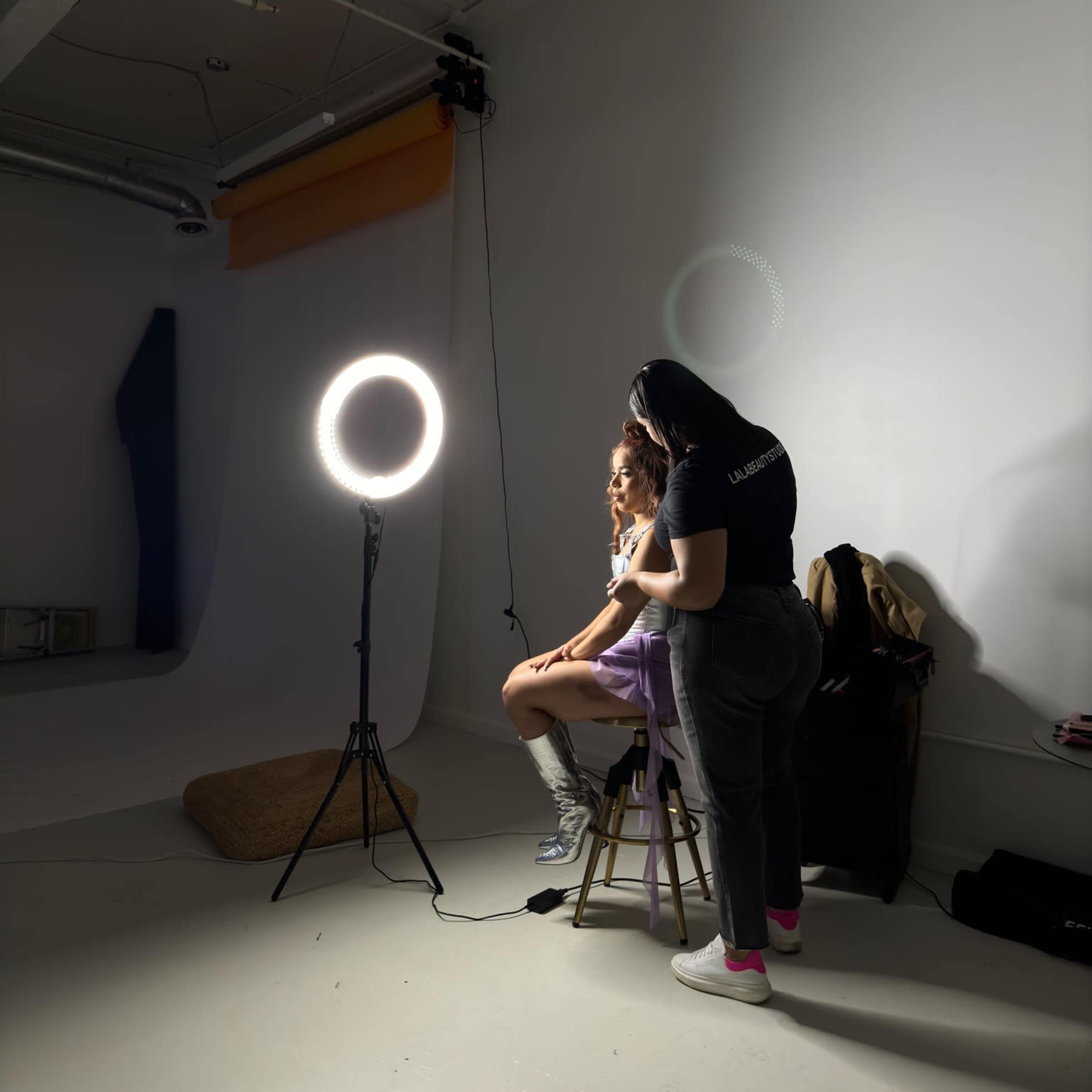 A model sits on a stool in a photography studio while a stylist adjusts her hair next to a ring light.